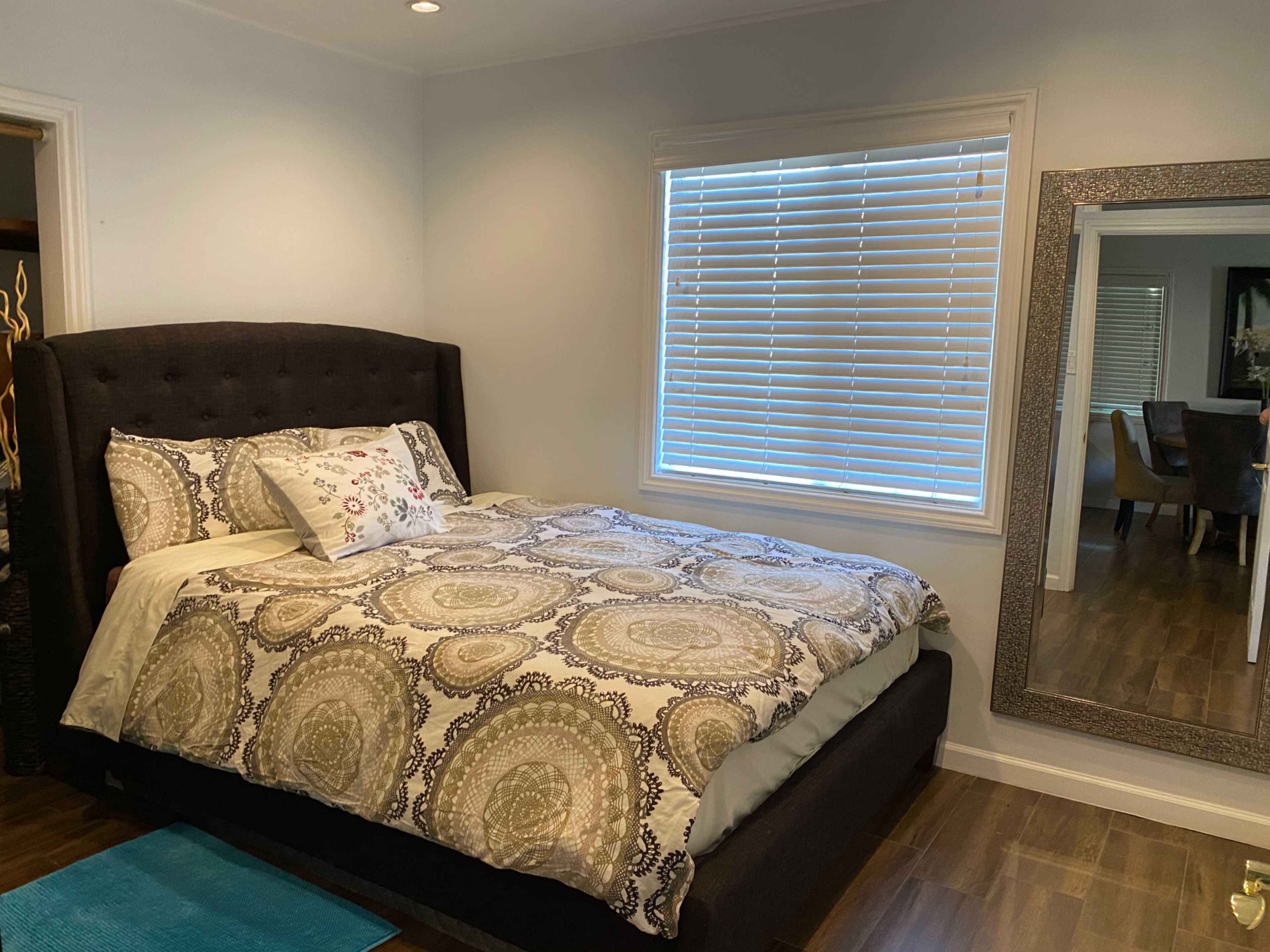 A neatly made bed with a patterned comforter is positioned against a wall in a well-lit bedroom, featuring a large mirror and a window with horizontal blinds.