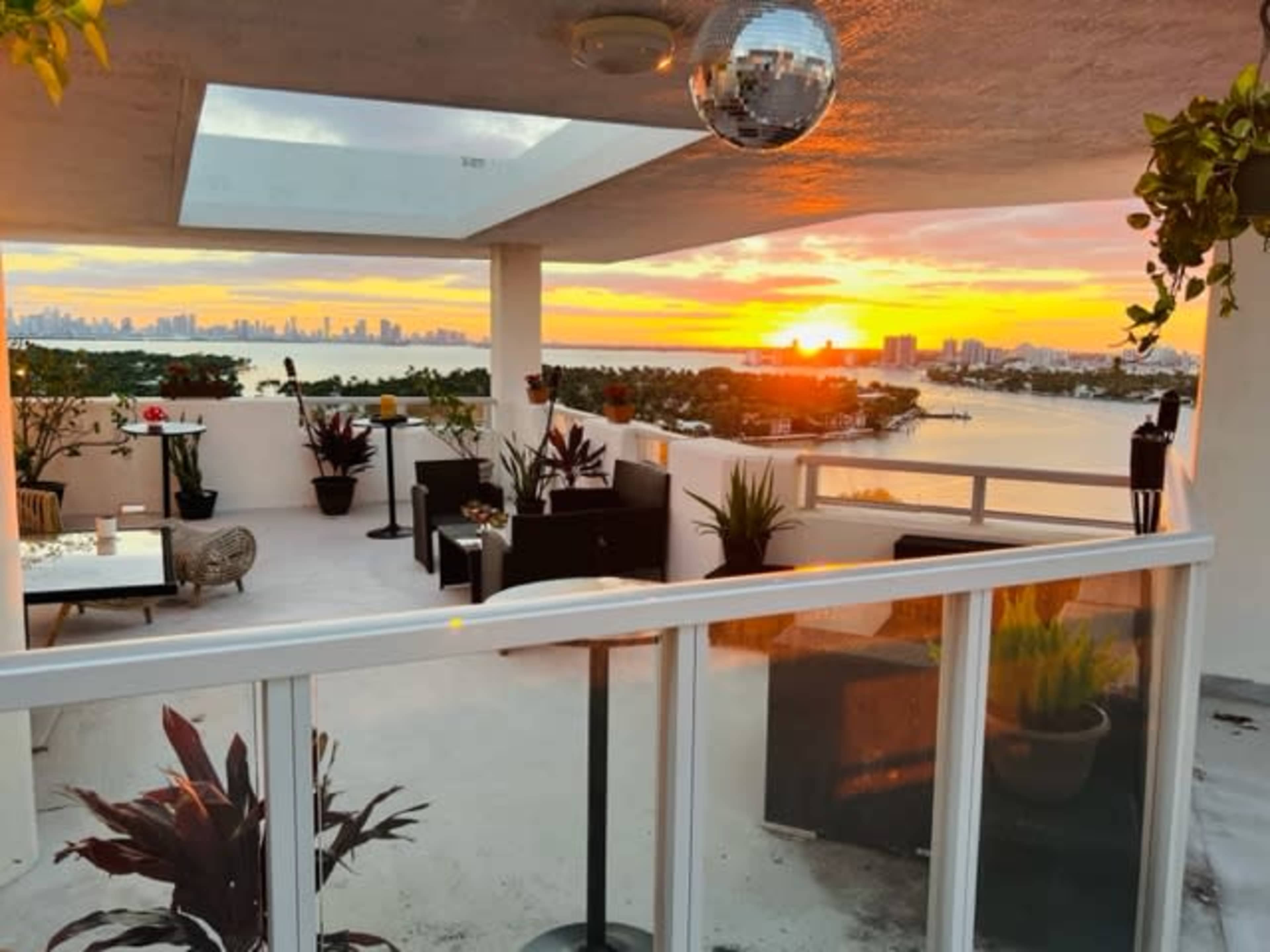 A rooftop patio adorned with potted plants features comfortable seating and a view of a sunset over the water and city skyline.