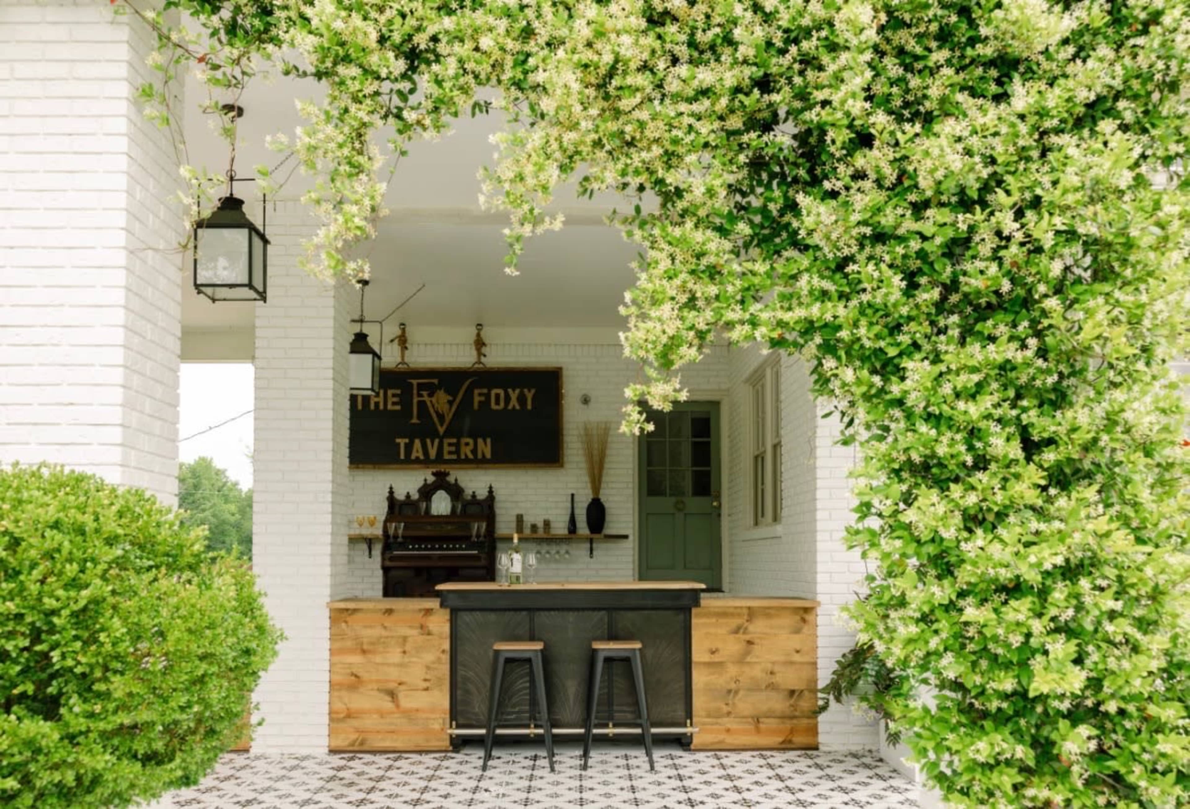The image shows an outdoor bar area with a black countertop, wooden stools, and a backdrop featuring a sign that reads "THE FOXY TAVERN," framed by lush greenery and flowering vines.