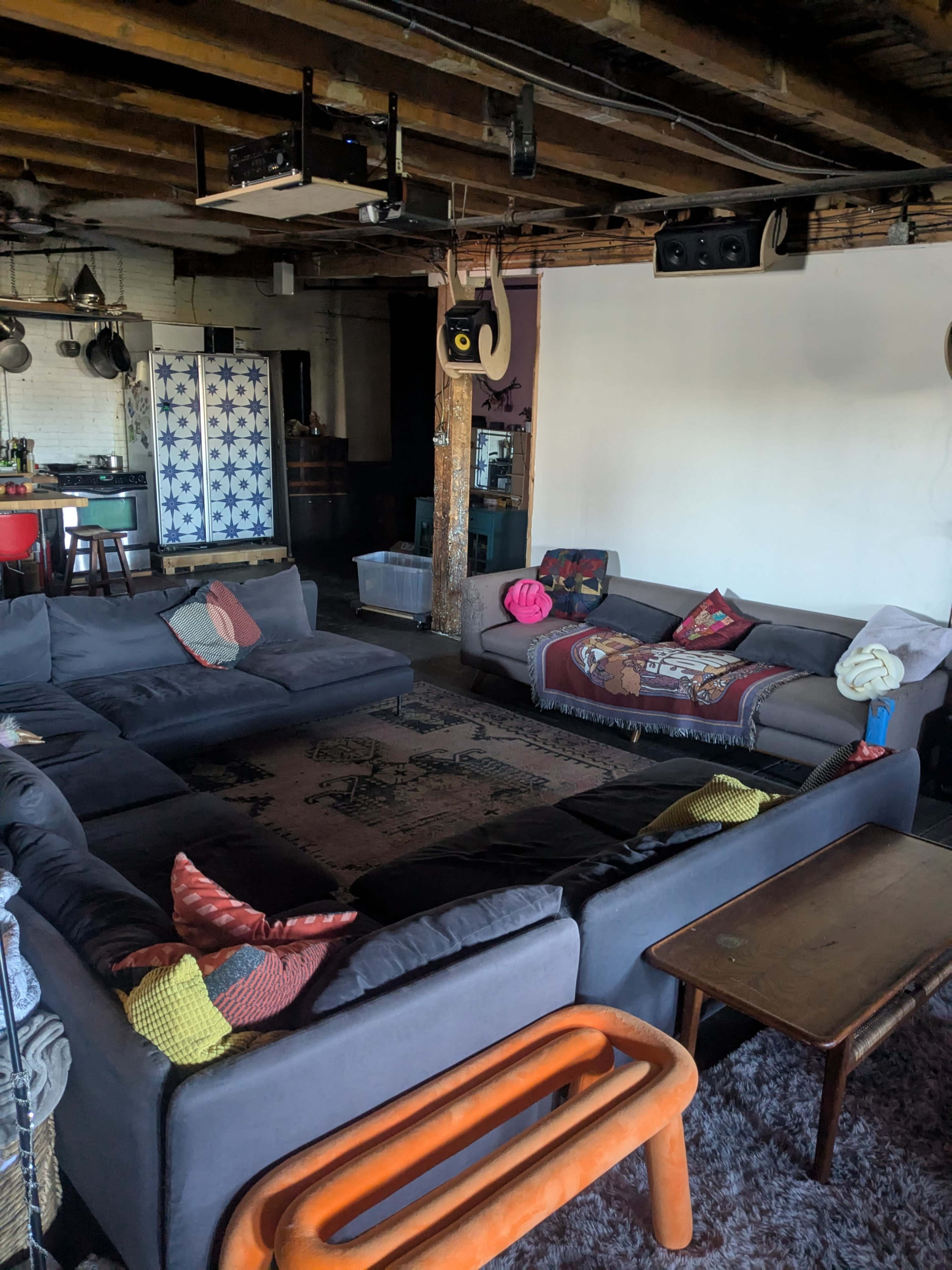 The image shows a spacious living area with two gray sofas arranged in a circular layout around a central rug, wooden beams on the ceiling, and various decorative items scattered around.