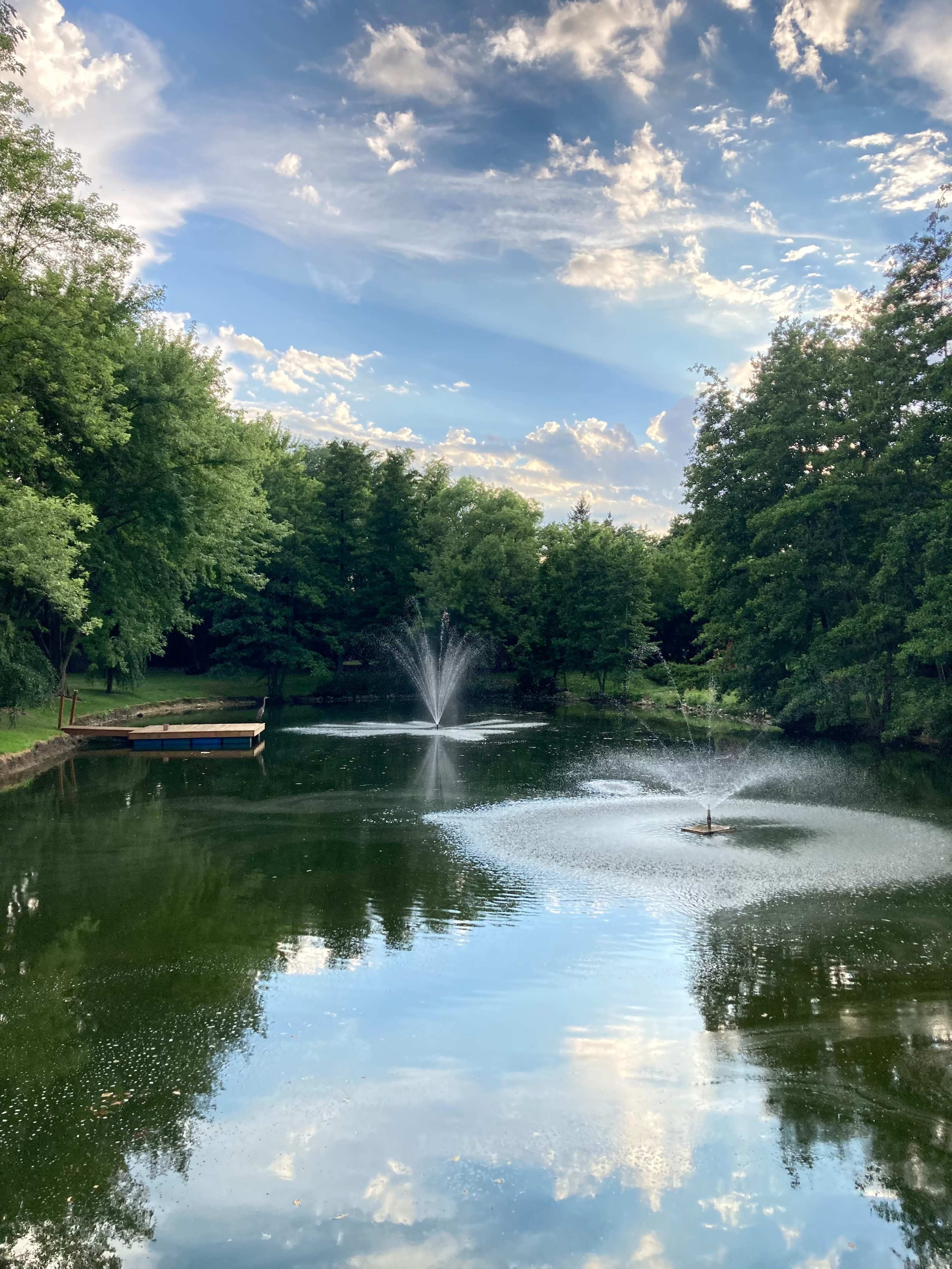 A tranquil pond surrounded by trees features two fountains, reflecting the sky and foliage in the calm water.