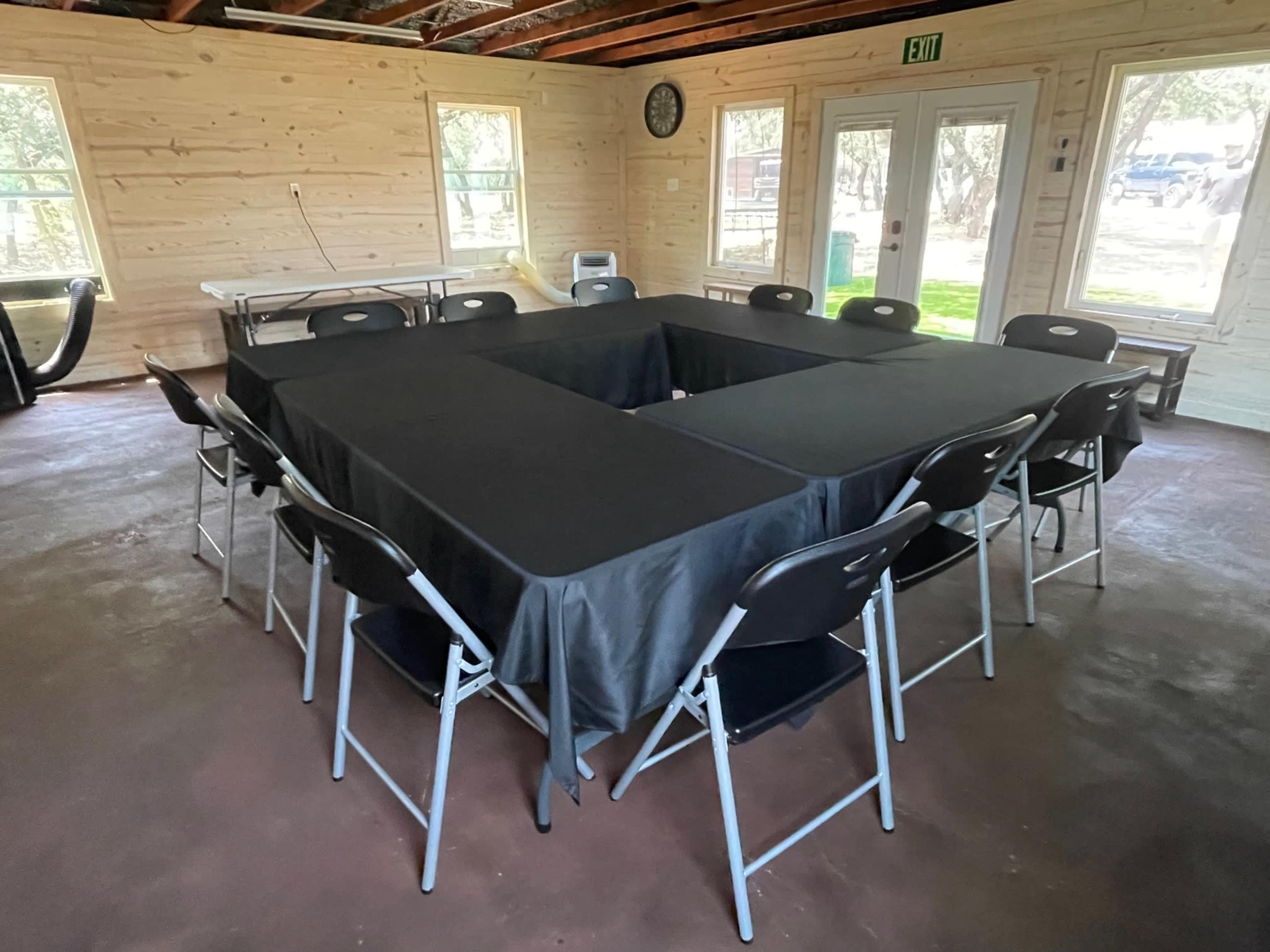 A square arrangement of tables covered with black cloth and surrounded by folding chairs is set up in a wooden interior space with large windows.
