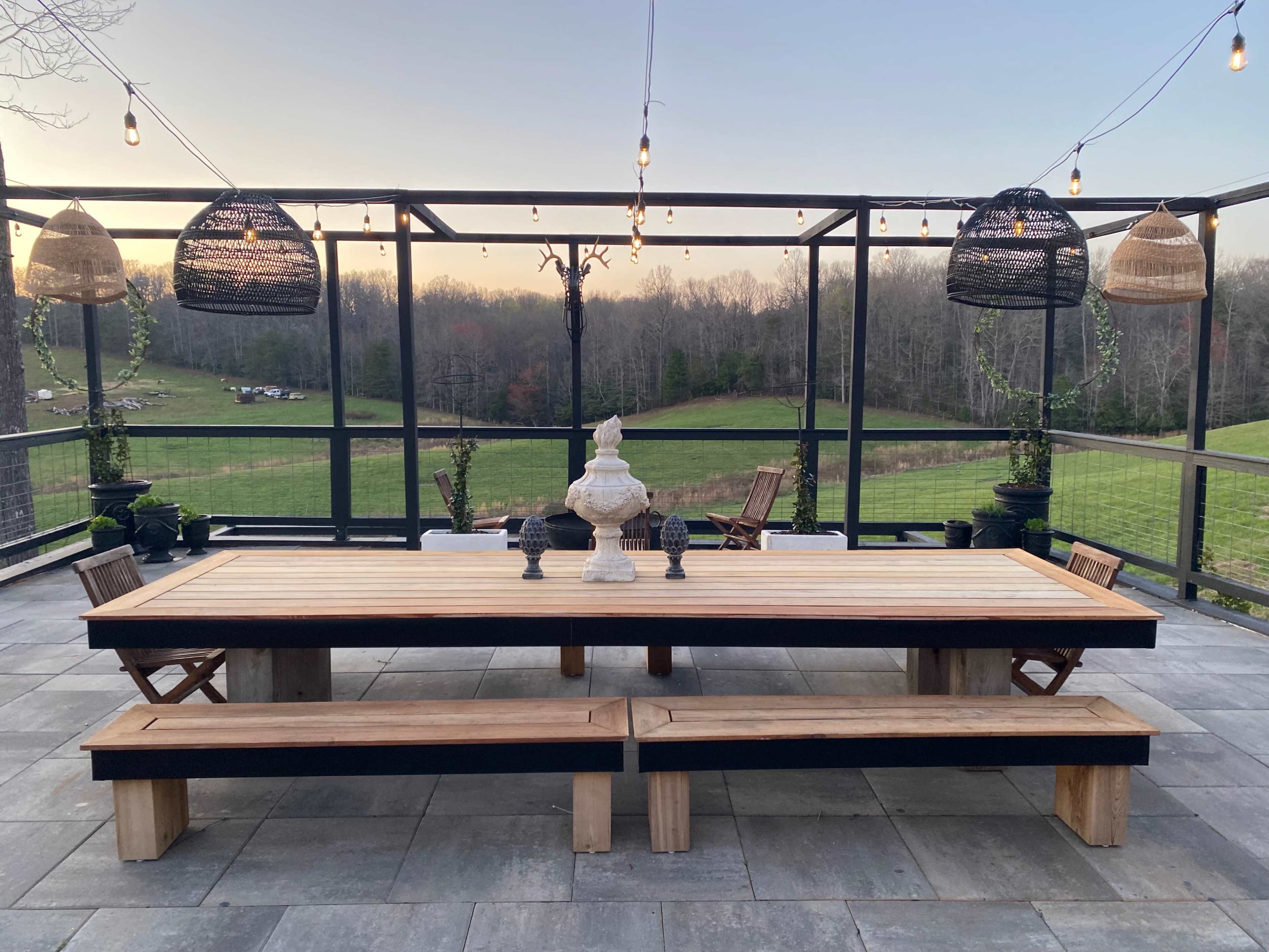 A spacious outdoor dining area features a large wooden table, benches, and decorative pendant lights, set against a backdrop of a green field and forested area.