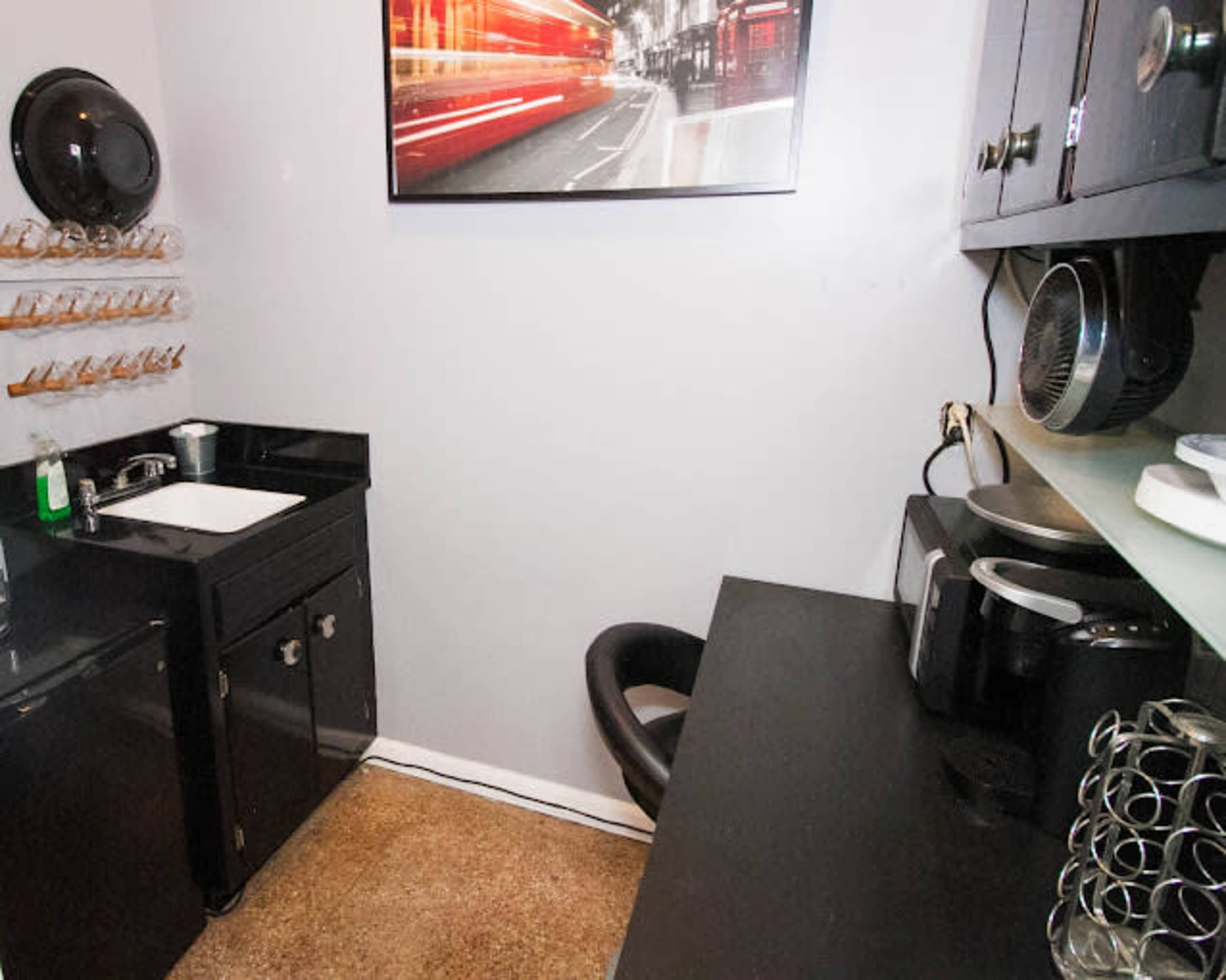 The image shows a compact kitchen space featuring a black countertop, a small sink, a coffee maker, and a chair in front of a wall-mounted photograph.