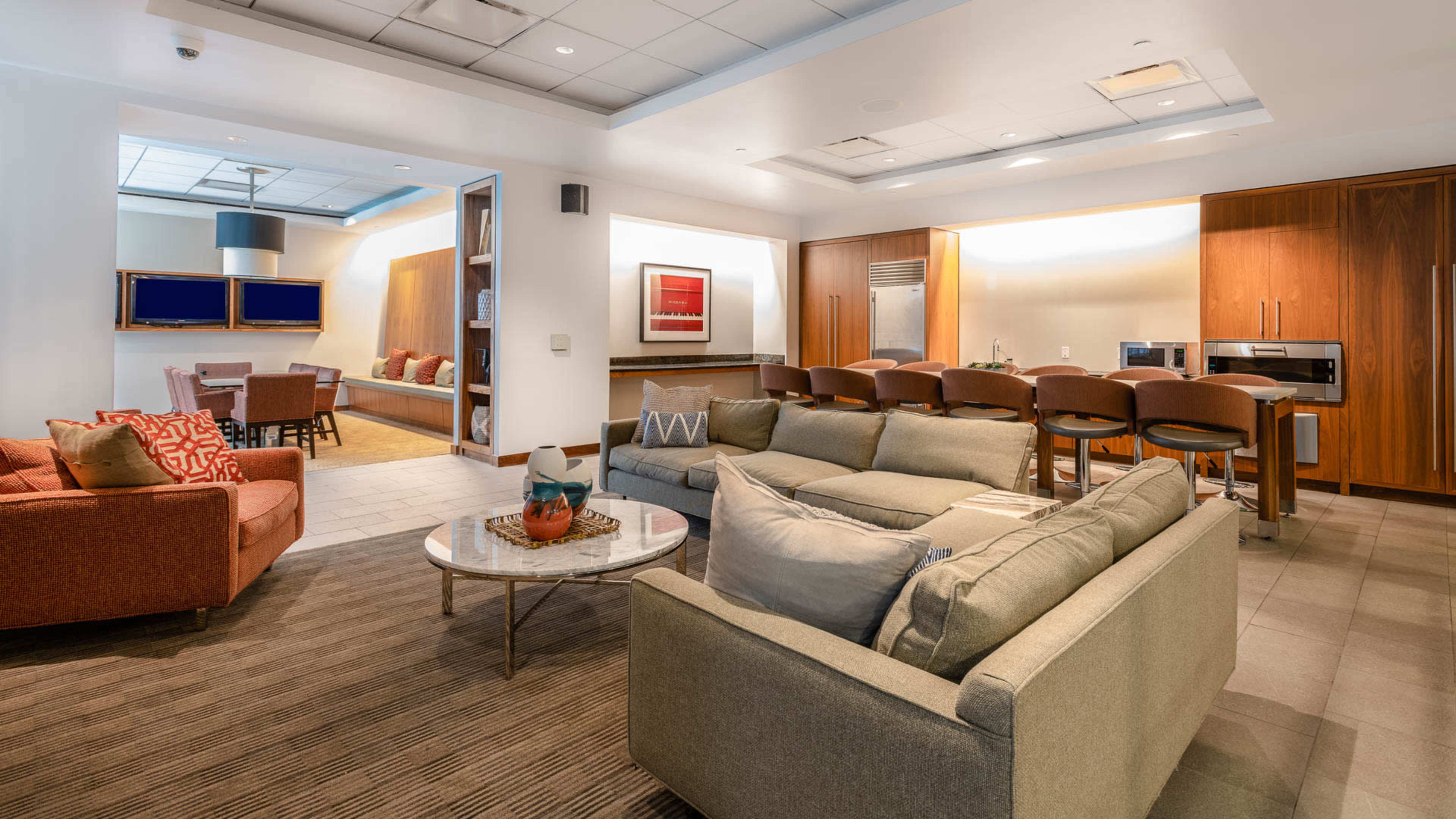 The image shows a modern lounge area featuring two sofas, a coffee table, and a kitchen with bar seating and appliances.