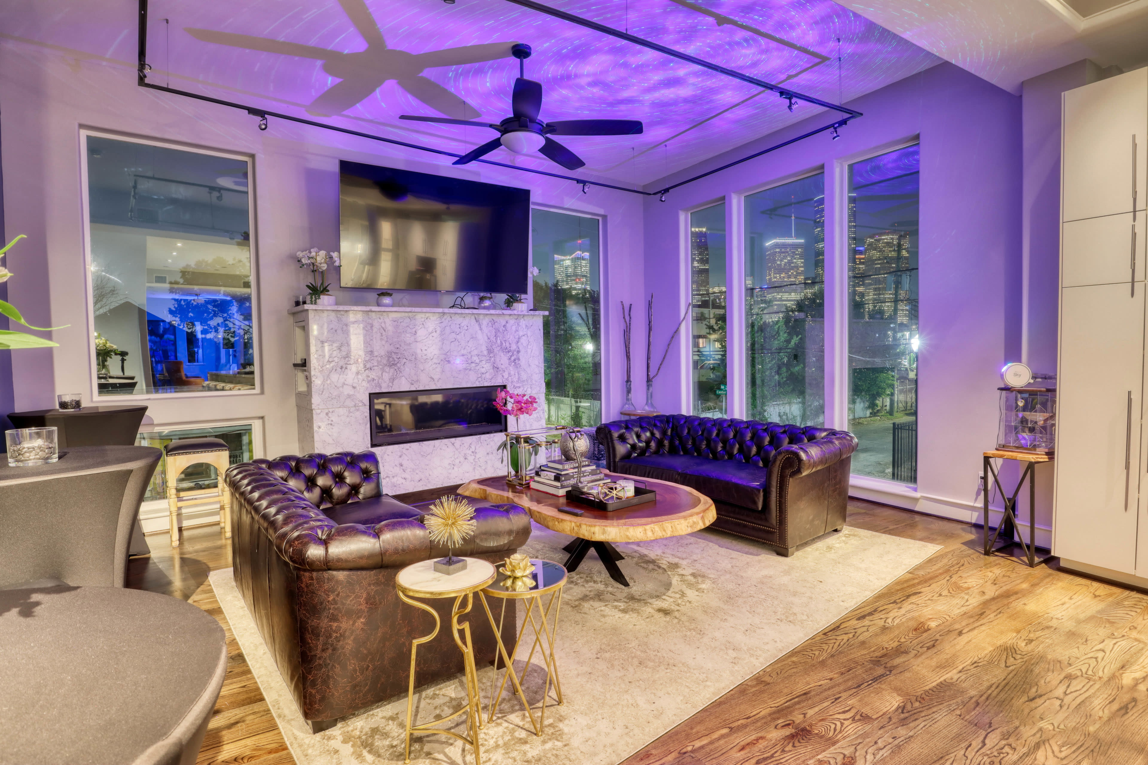 A modern living room with a marble fireplace, two leather sofas, a round coffee table, and large windows offering a view of a city skyline.