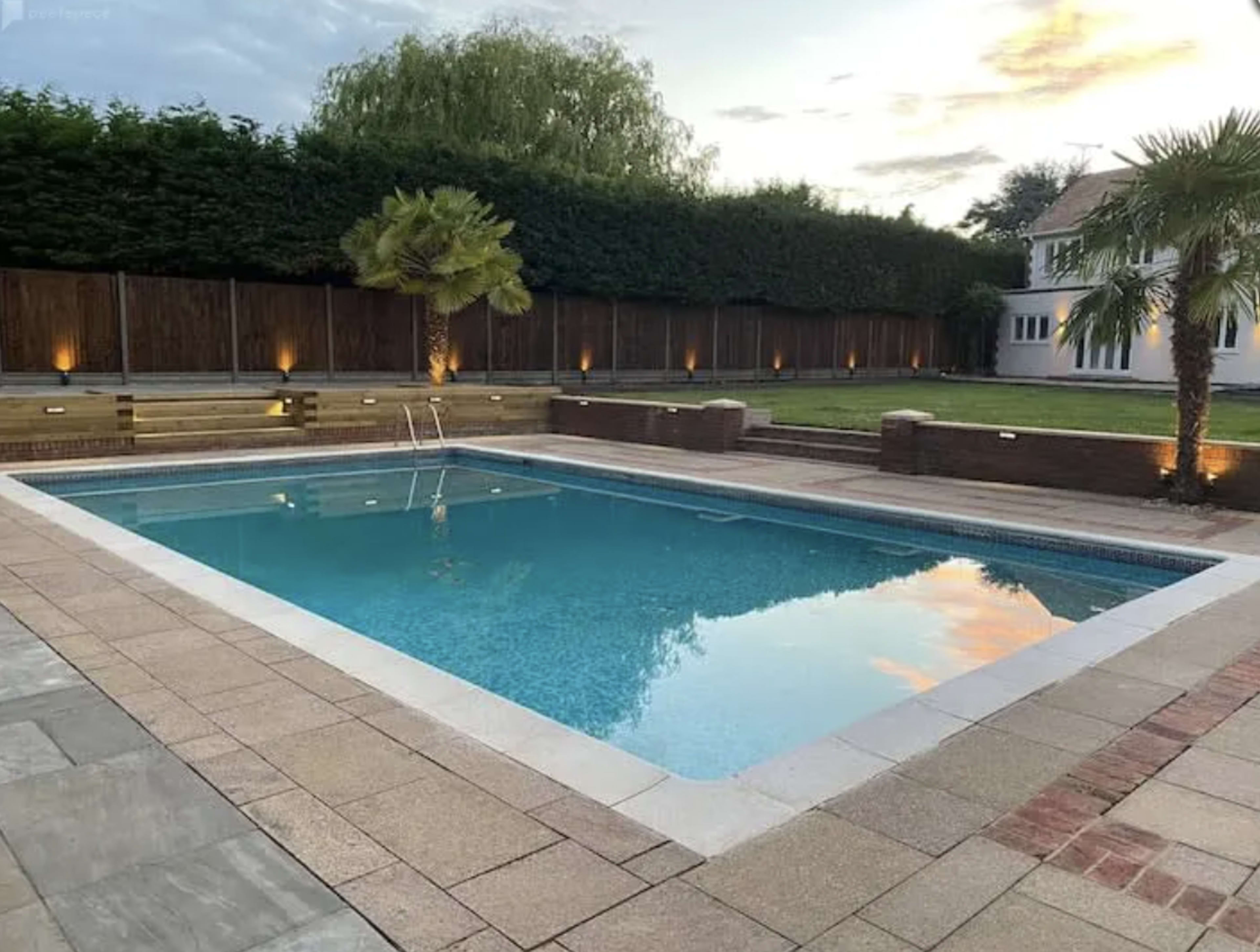 A rectangular swimming pool surrounded by paved stones, with palm trees and a well-maintained lawn in the background.