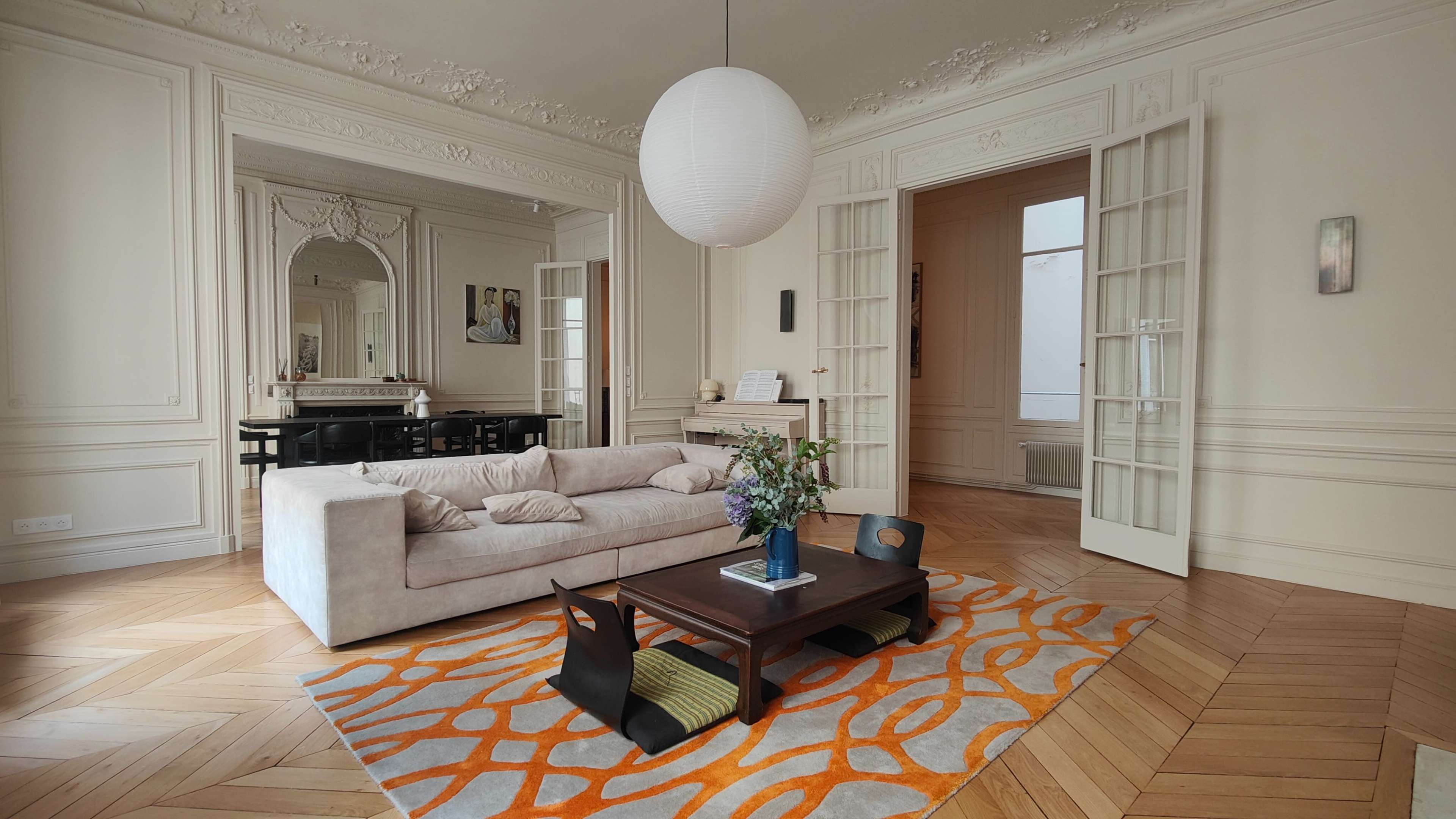 The image shows a bright living space featuring a light-colored couch, a wooden coffee table on an orange patterned rug, and a large round pendant light, with a mirror and another room visible in the background.