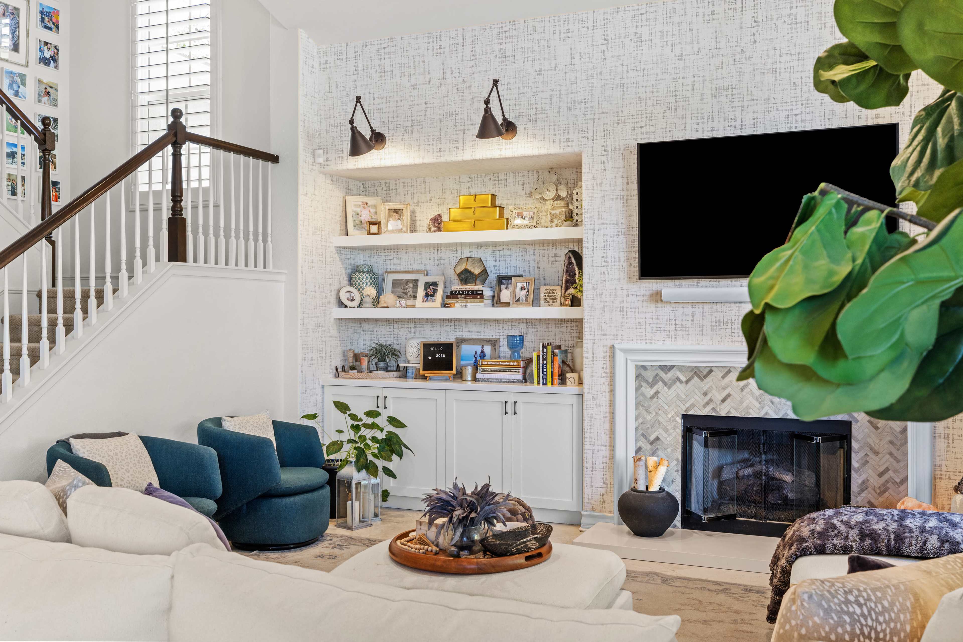 The image shows a modern living room featuring a cozy seating area with blue chairs, a built-in shelf with decorative items, and a fireplace beneath a flat-screen TV.