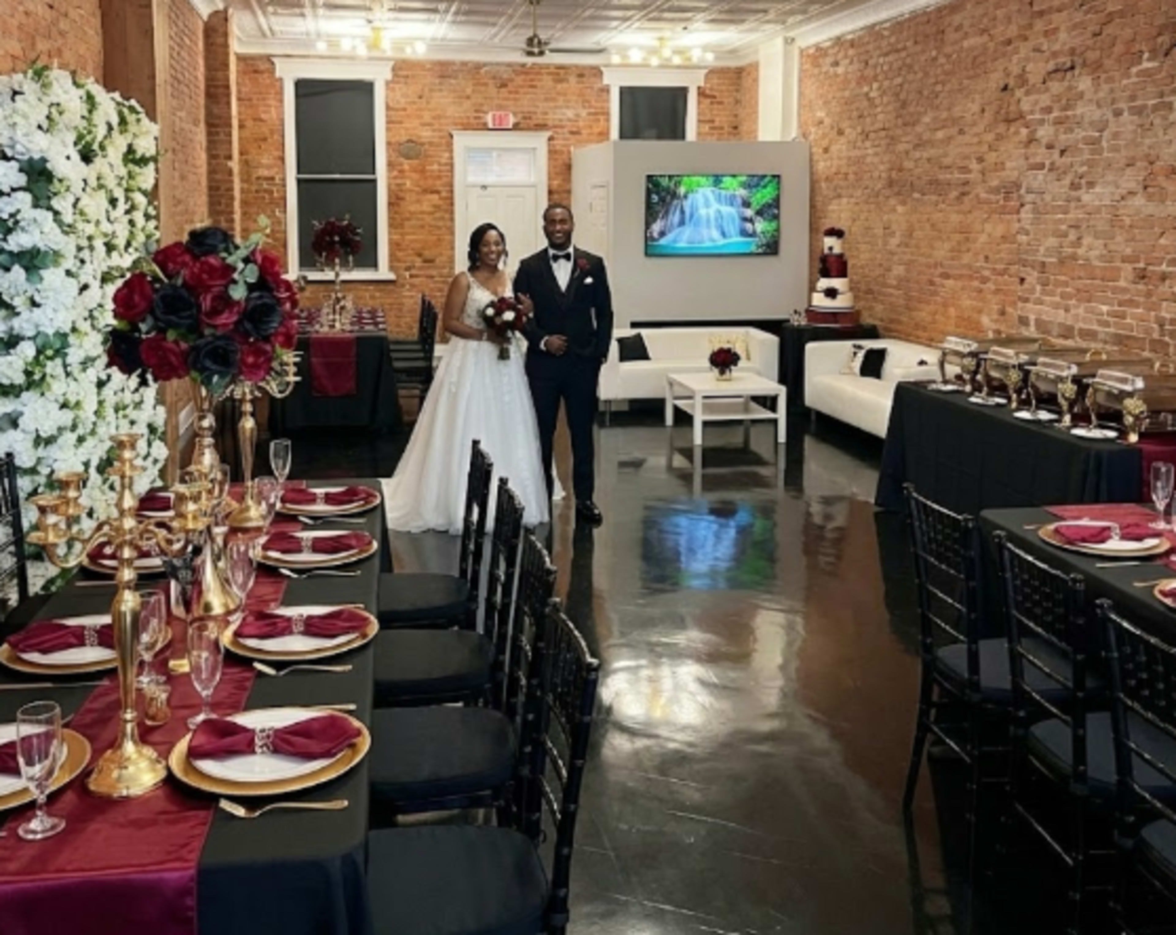 A couple stands in a decorated event space with exposed brick walls, featuring tables set for a formal gathering.