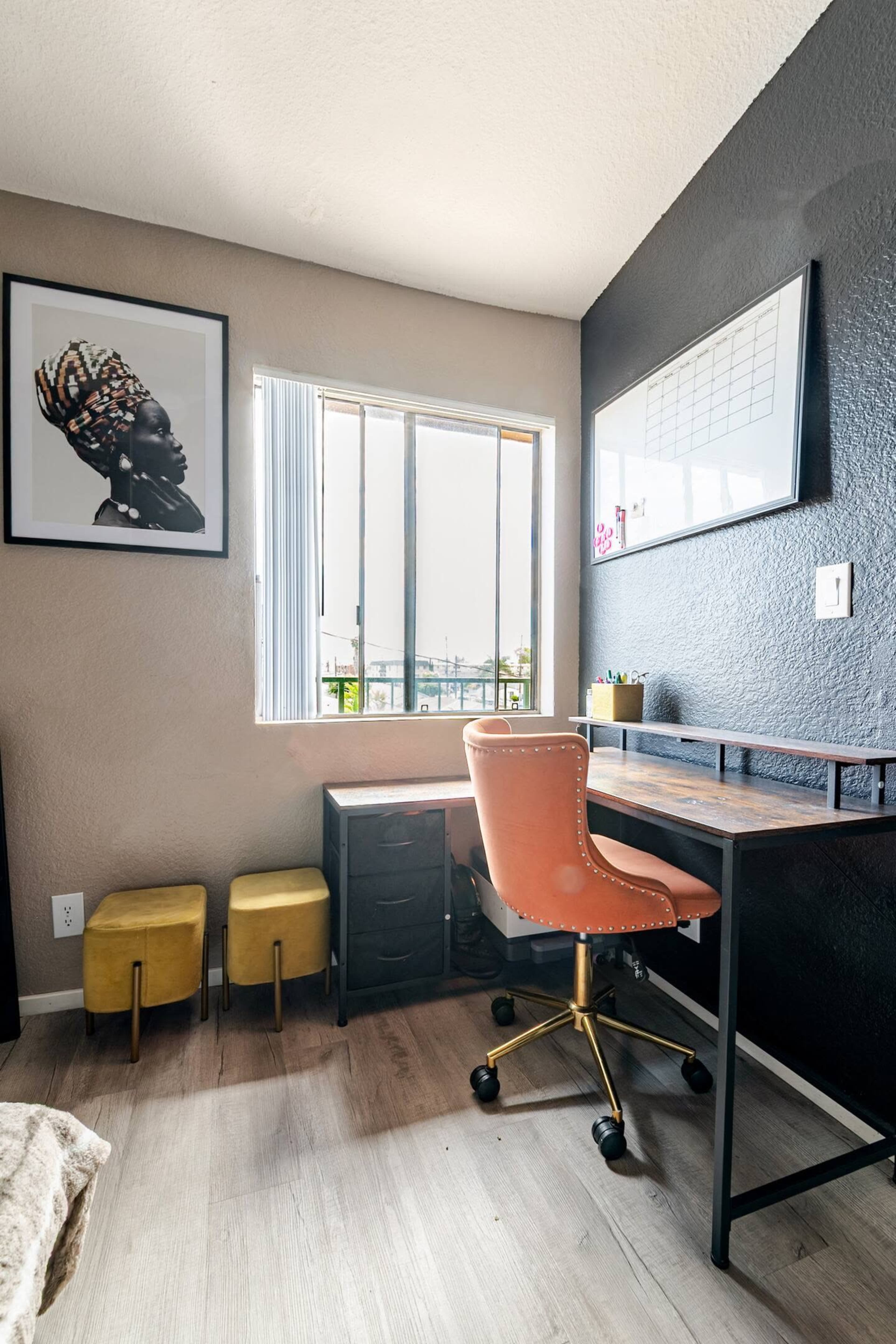The image shows a small workspace featuring a wooden desk with a chair, a calendar on the wall, and a window that lets in natural light, along with two yellow stools nearby.