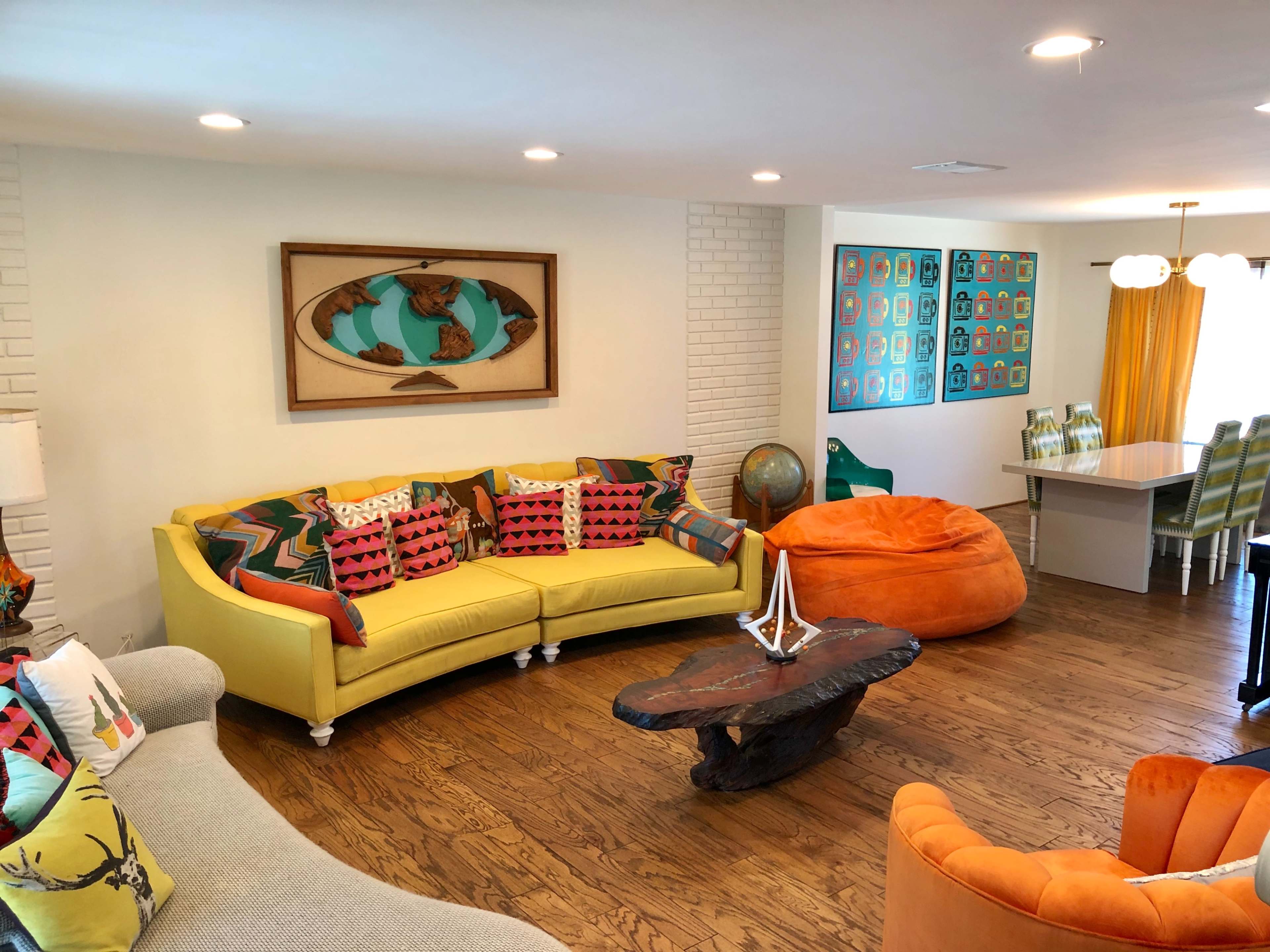 The living room features a yellow sectional sofa, various colorful cushions, a wooden coffee table, and a dining area with a large table and patterned chairs.