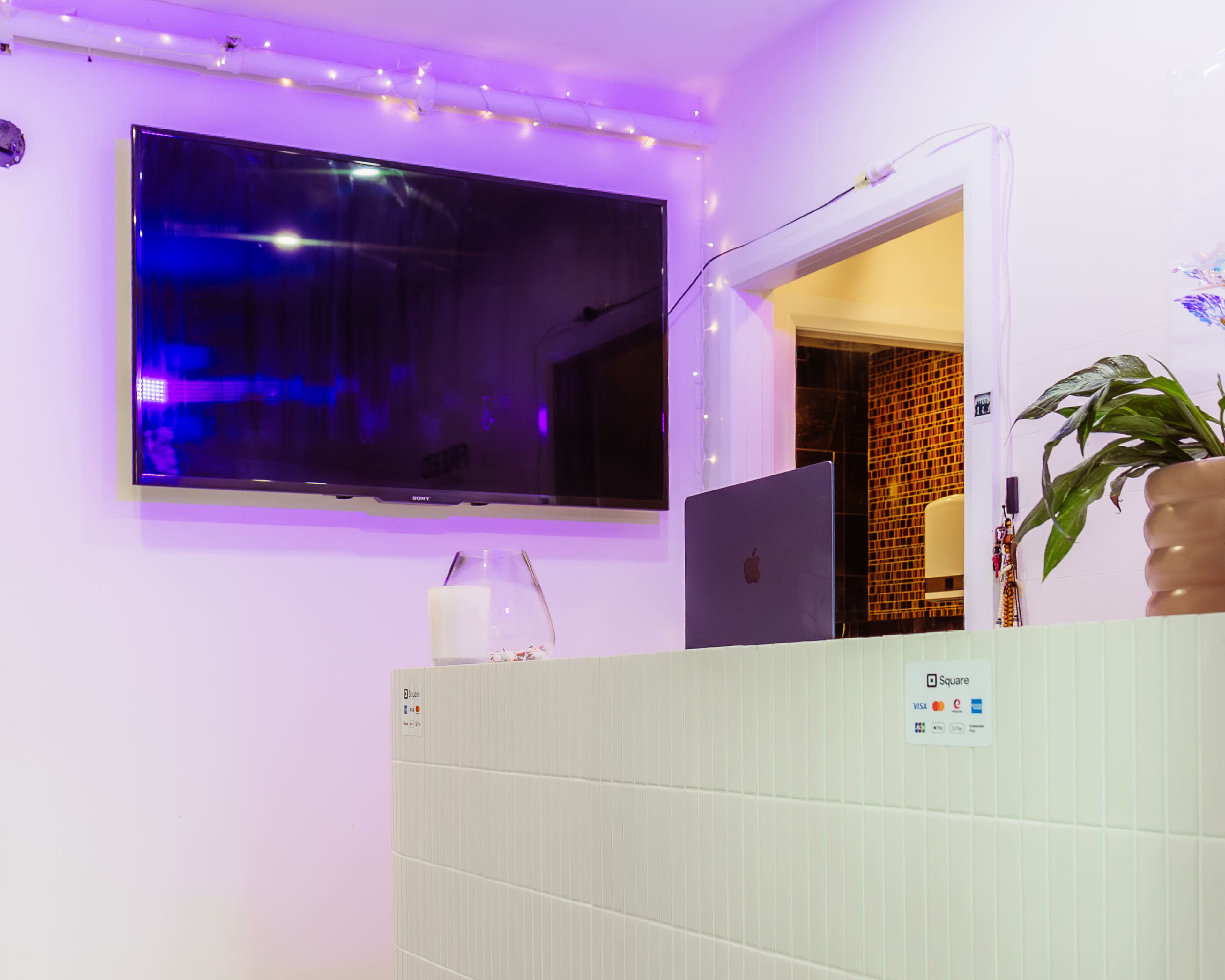 A modern reception area features a sleek white counter with a laptop and a large wall-mounted television above, illuminated by soft purple lighting.
