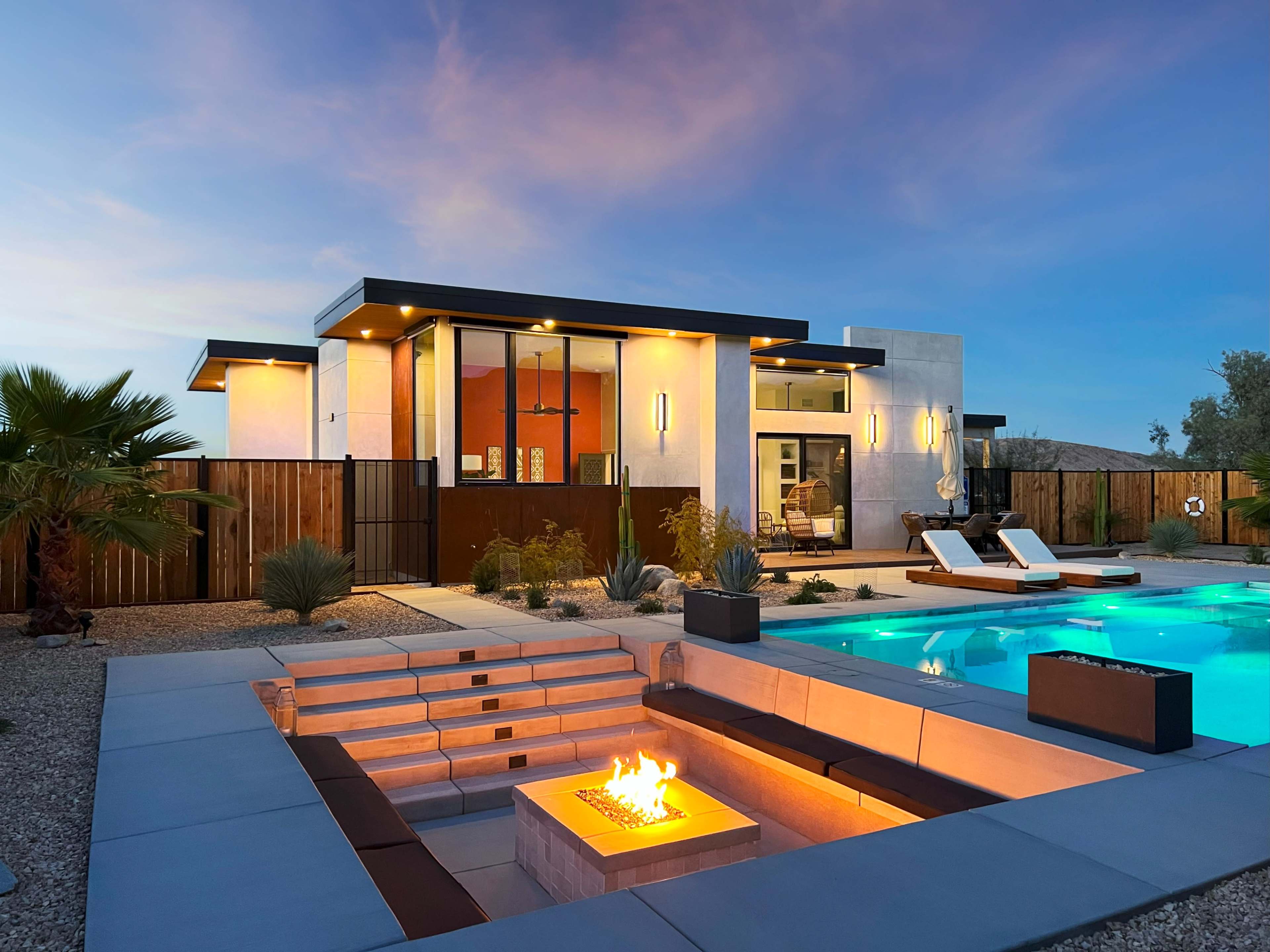 The image depicts a modern home with a swimming pool and a fire pit in the foreground, surrounded by desert landscaping under a twilight sky.