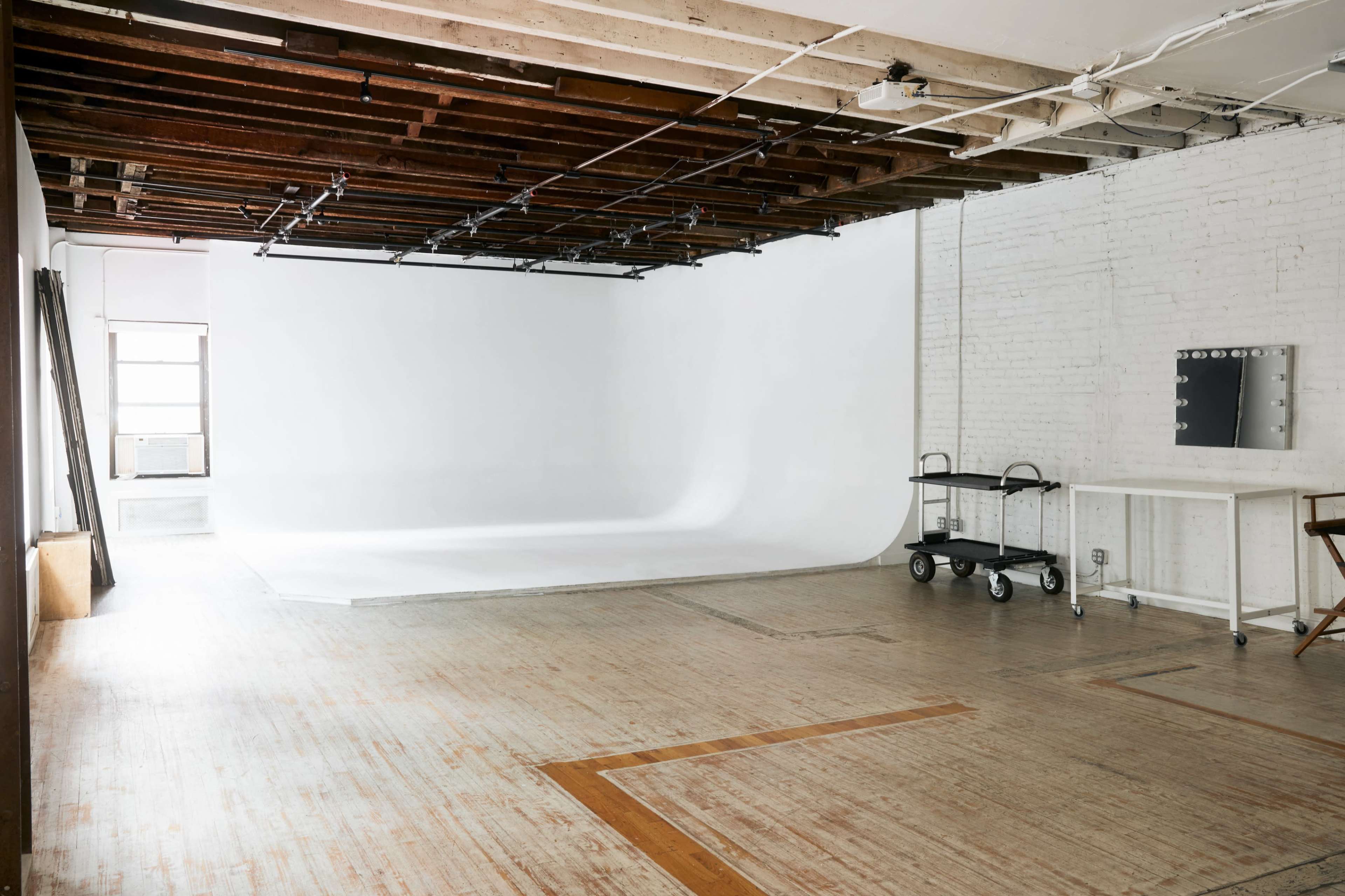 A spacious, empty photography studio with a white backdrop, wooden floors, and a makeup station on the side.