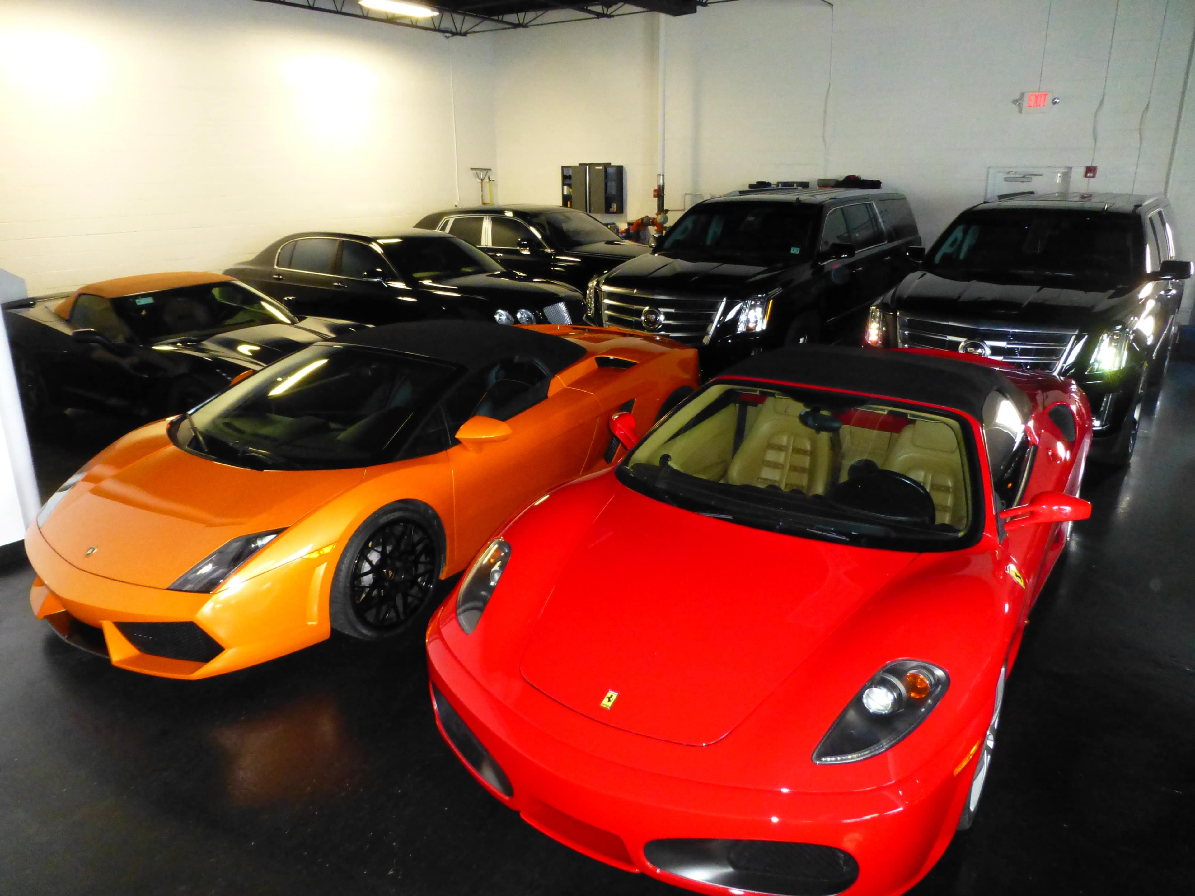 A collection of luxury cars, including an orange Lamborghini and a red Ferrari, is parked inside a dimly lit garage alongside several black SUVs.