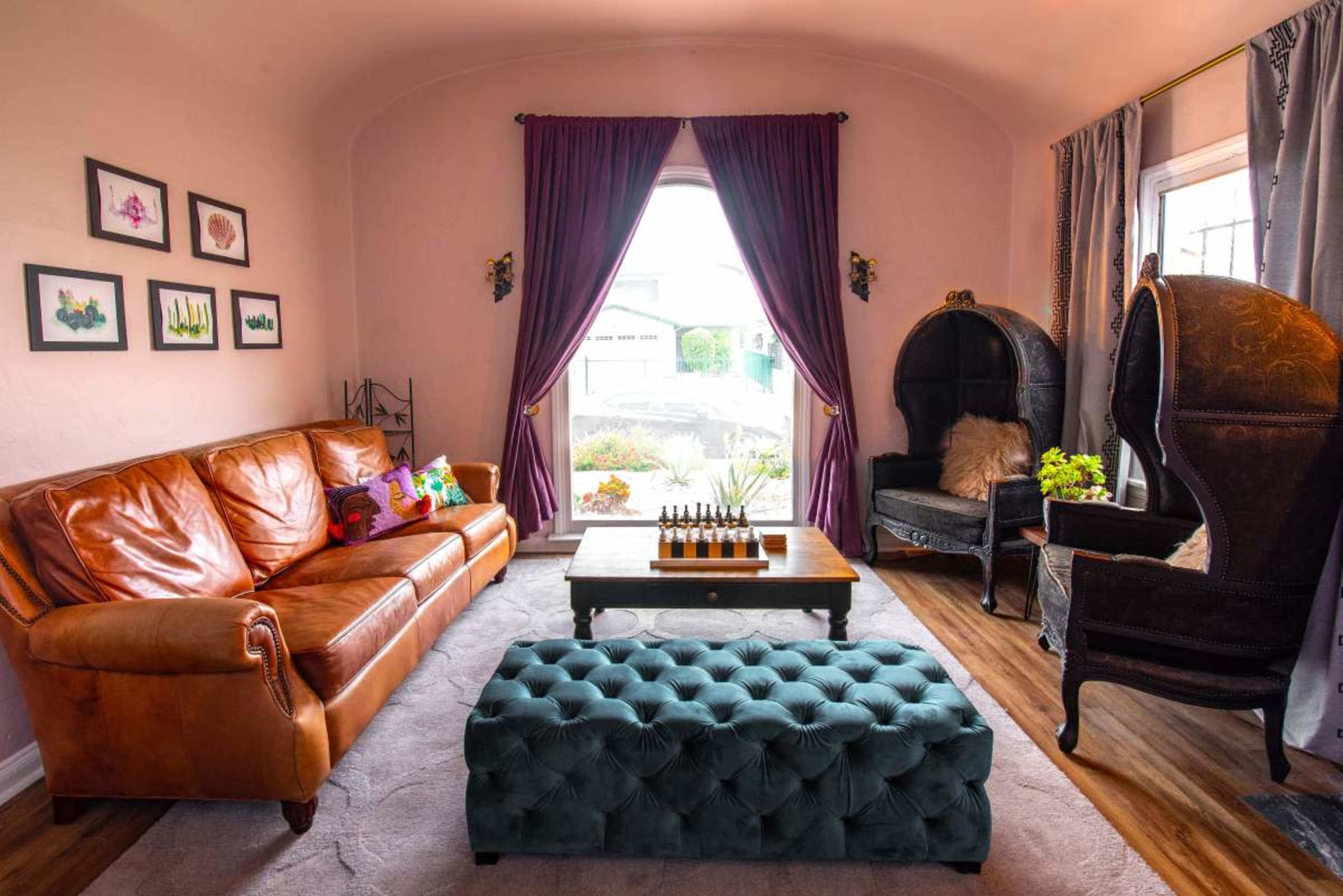 The living room features a brown leather sofa, a tufted teal ottoman, and two ornate high-backed chairs, with large windows allowing natural light to illuminate the space.