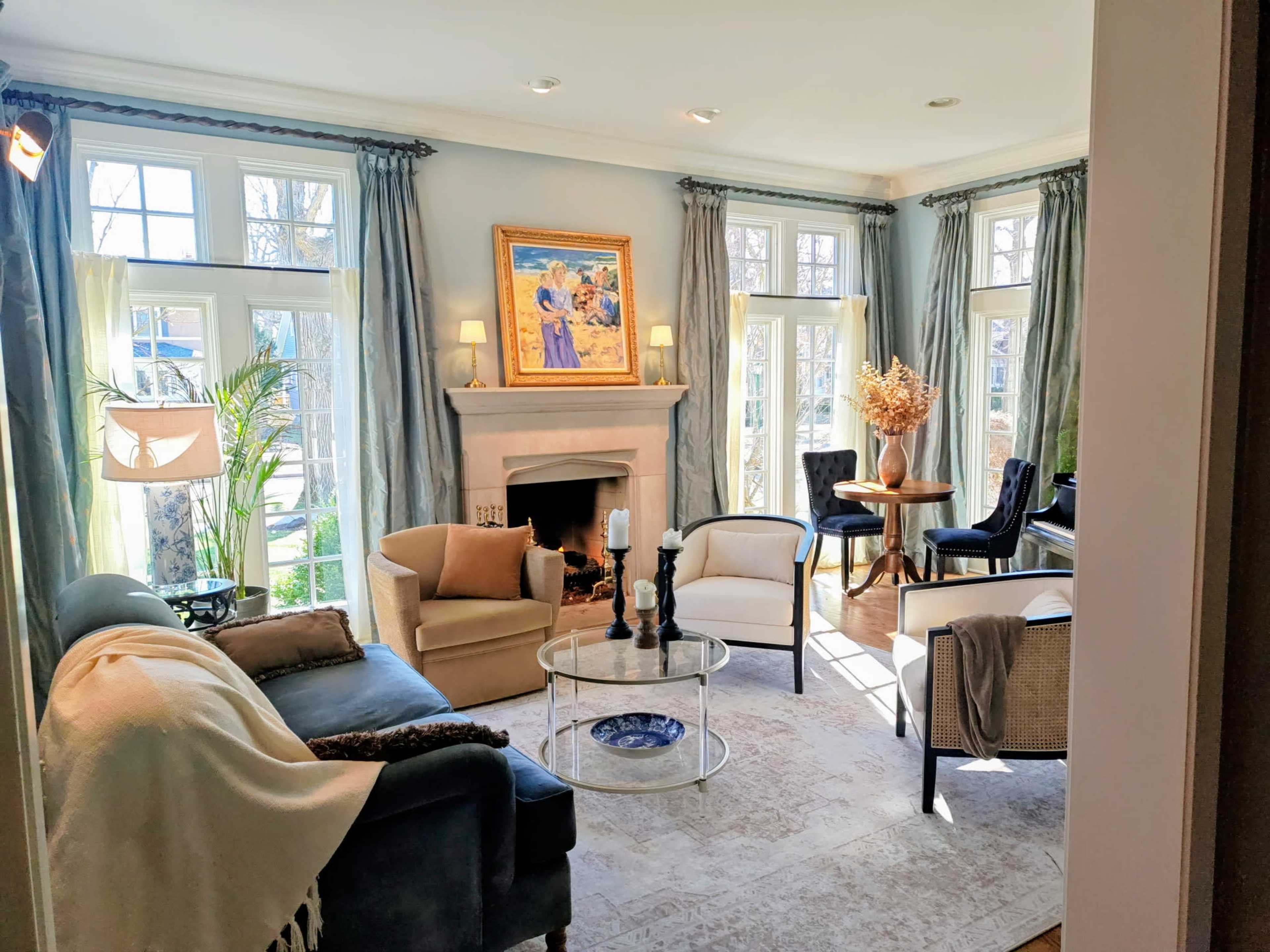 A modern living room features large windows, a fireplace, a framed artwork, and a variety of seating options.