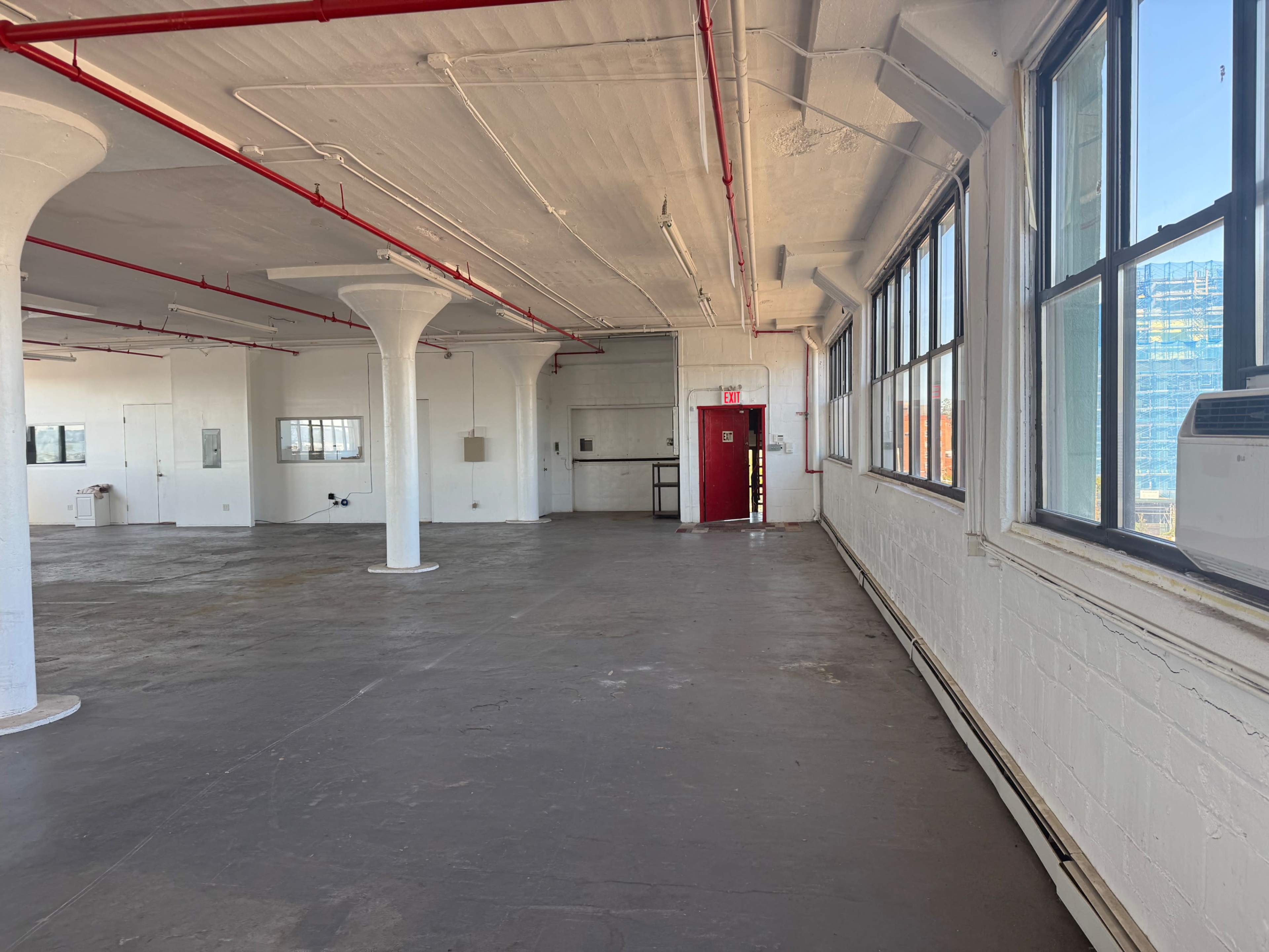 The image shows an empty industrial space with concrete floors, large windows, and exposed red piping along the ceiling.