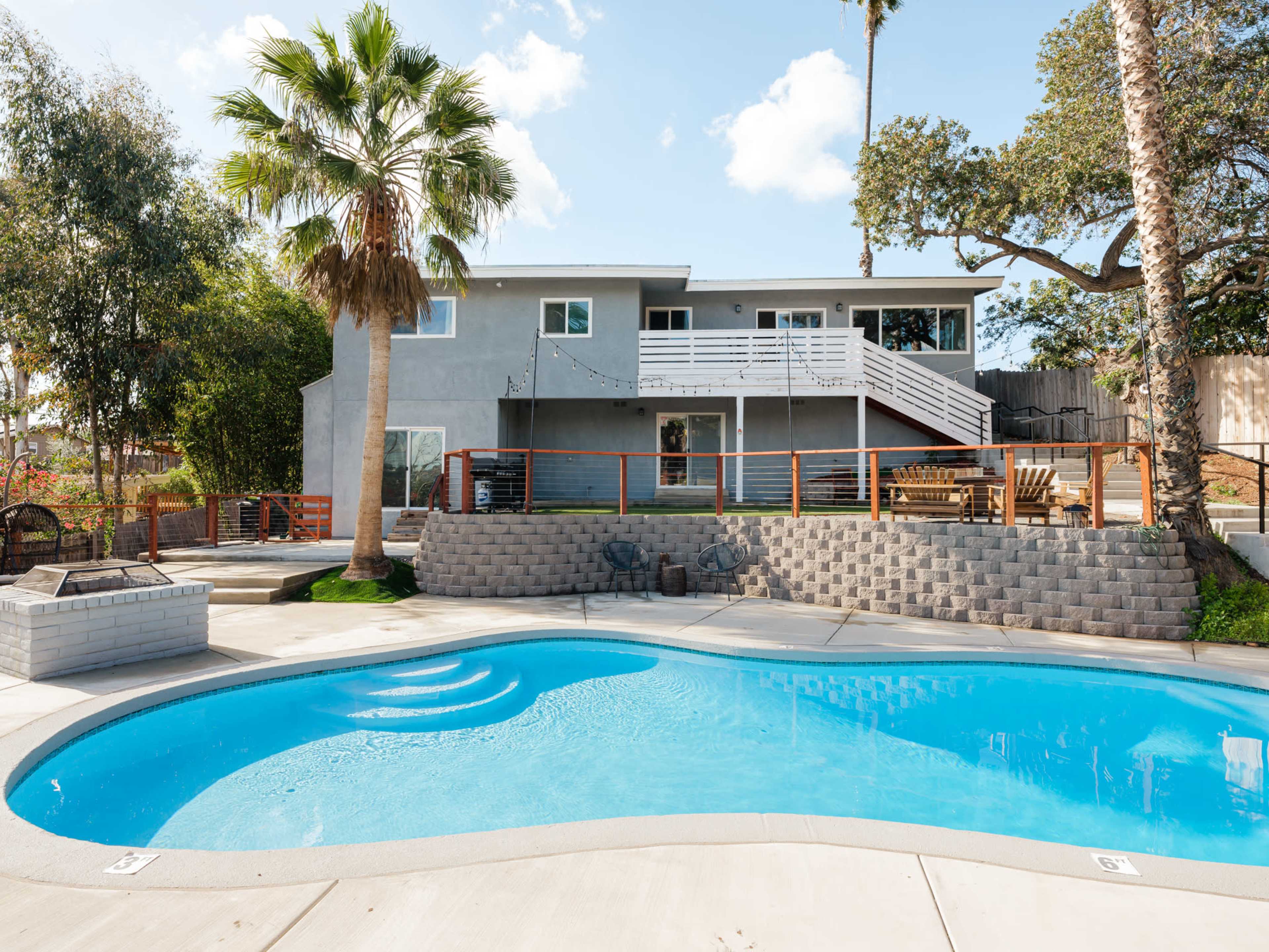A backyard features a blue swimming pool with a patio area and a two-story gray house in the background.