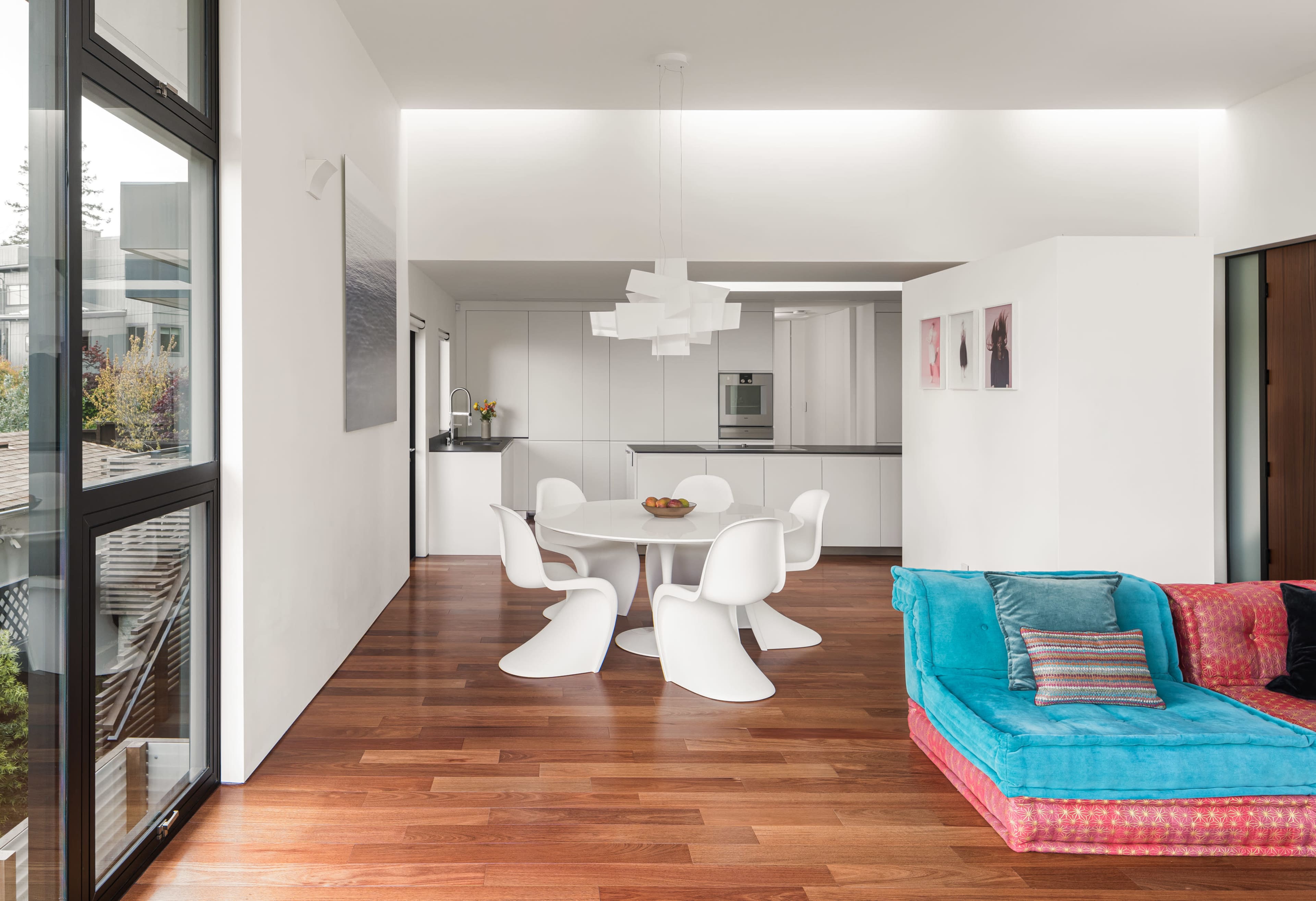 A modern, open-concept living space featuring a white dining table with chairs, a bright blue and pink sofa, wooden flooring, and minimalist kitchen cabinetry in the background.
