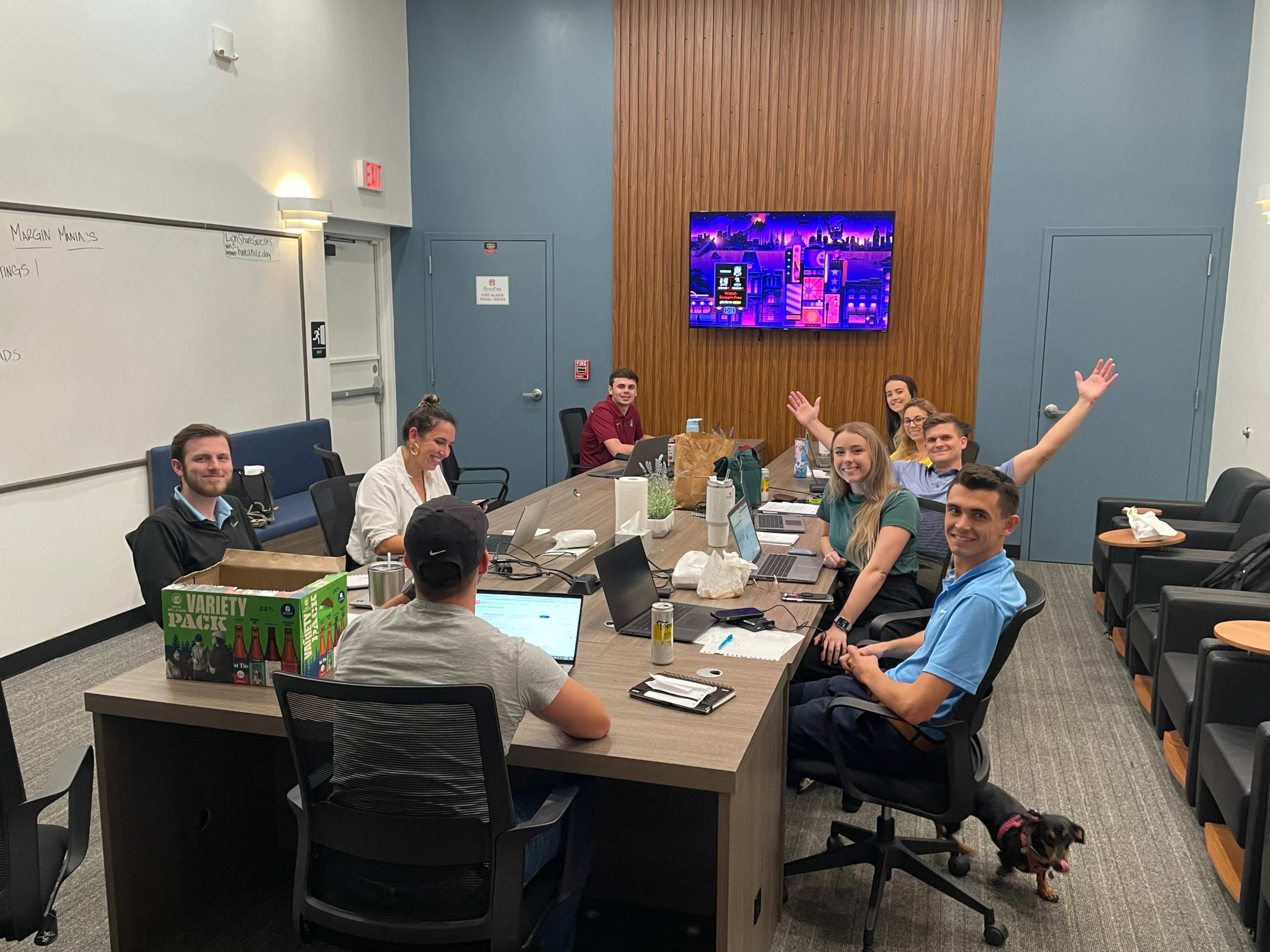 A group of eight people is seated around a conference table in a meeting room with laptops and a large screen displaying colorful content in the background.