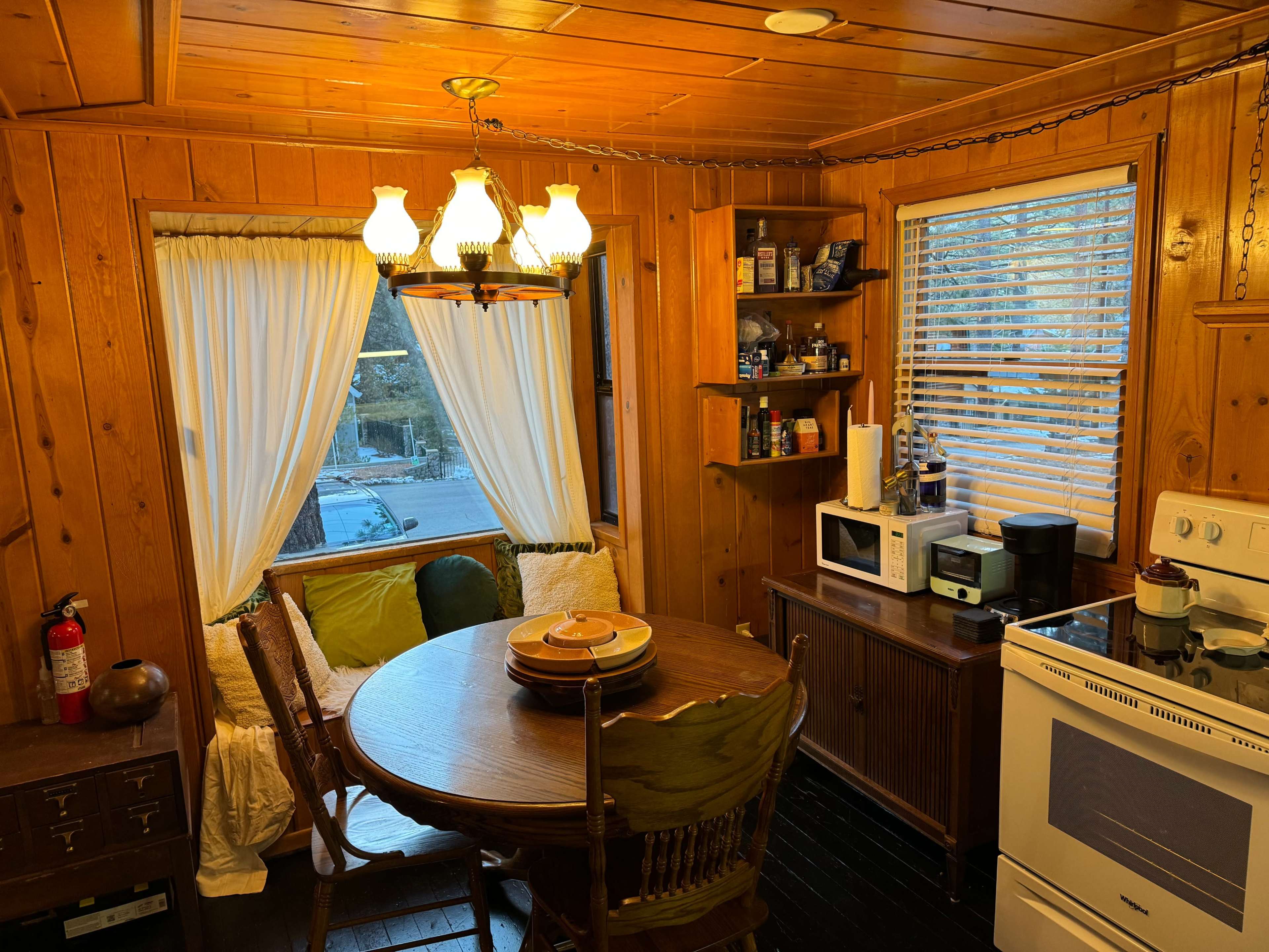 A cozy kitchen featuring a wooden table with chairs, a stove, a microwave, and a window with curtains, illuminated by a chandelier.