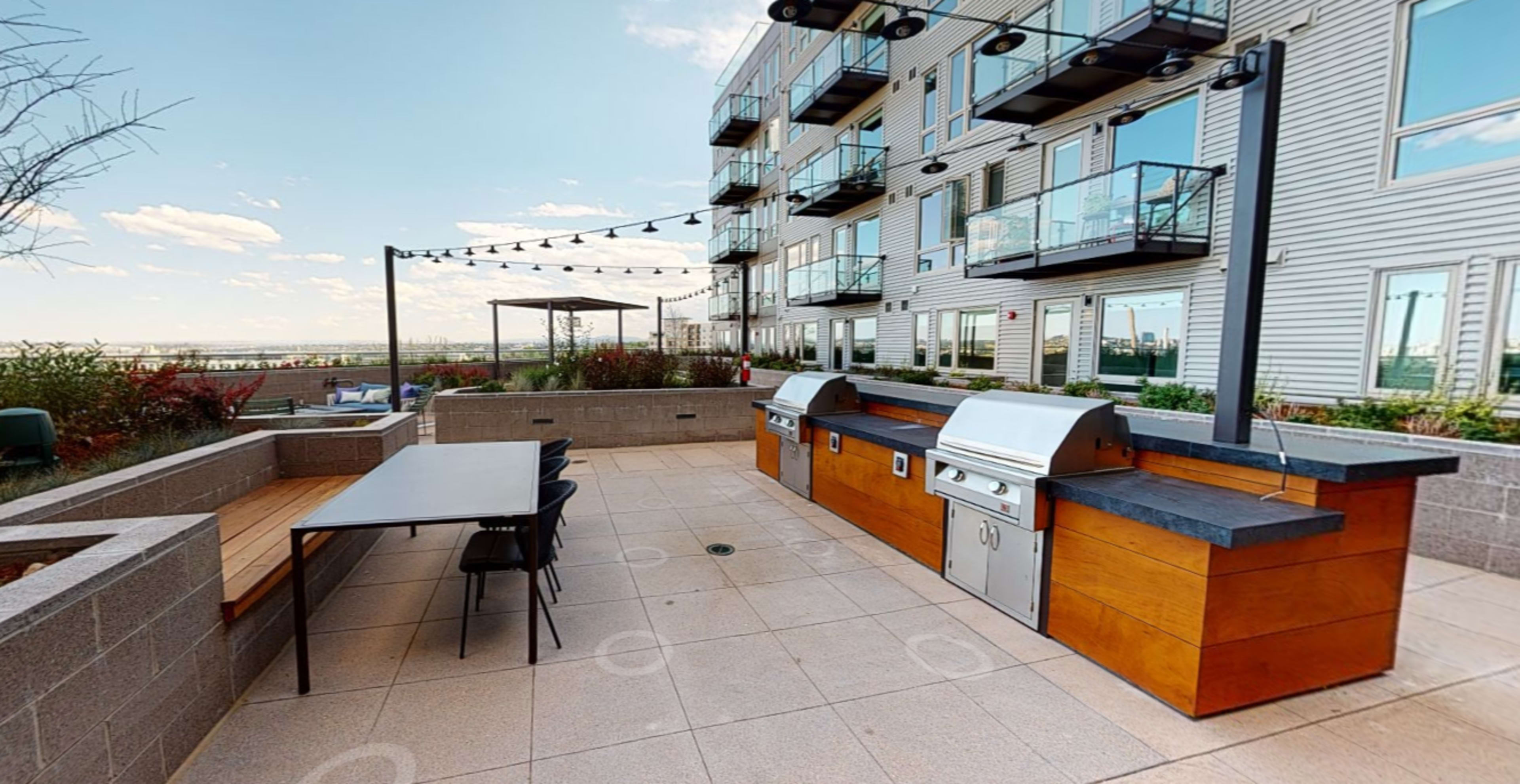 An outdoor patio area featuring a grill station, a long dining table, and several balconies of an apartment building in the background.
