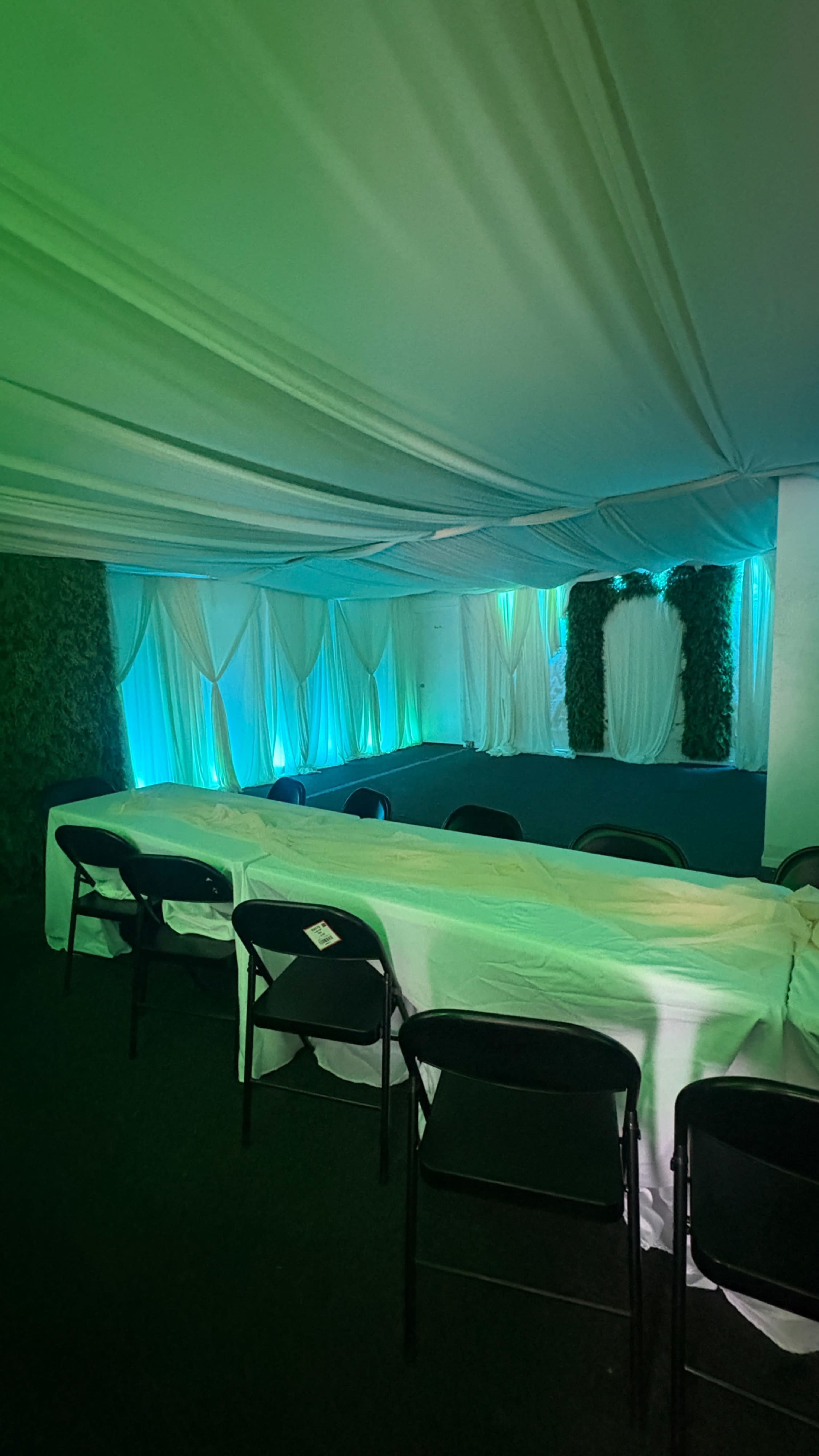 The interior of a tent is set up with a long table covered in a white cloth, surrounded by black chairs, and illuminated by soft green lighting.