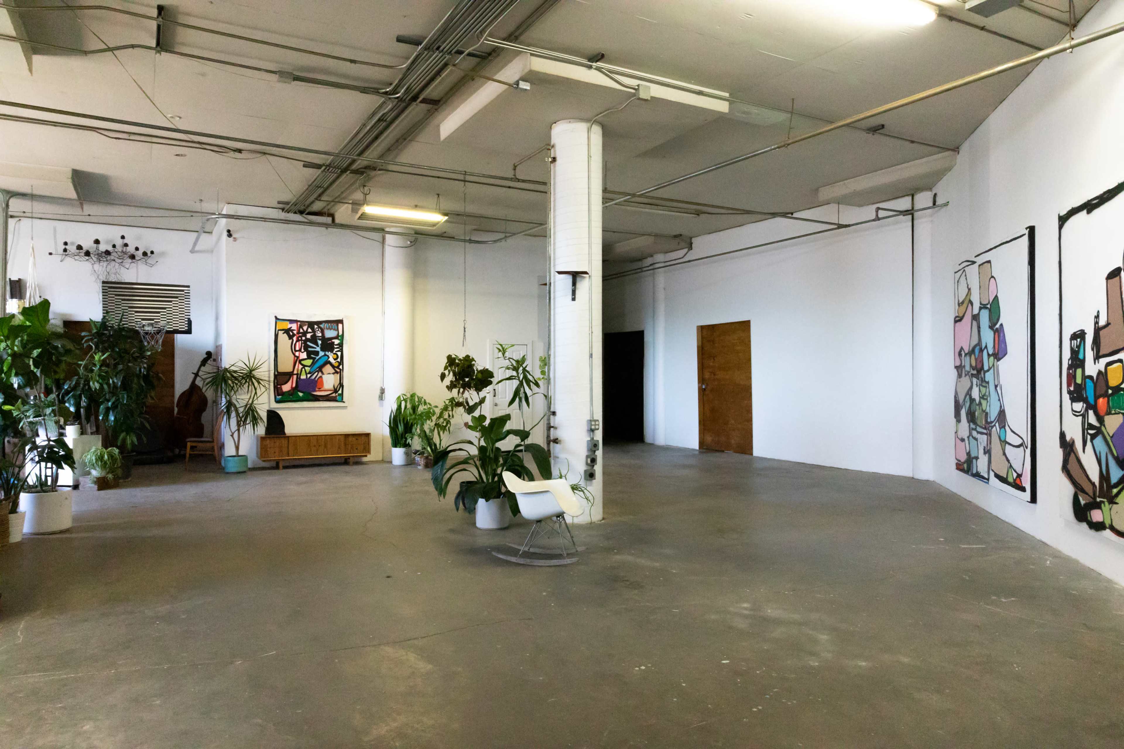 An expansive, empty interior space featuring artwork on the walls, potted plants, and minimal furnishings.