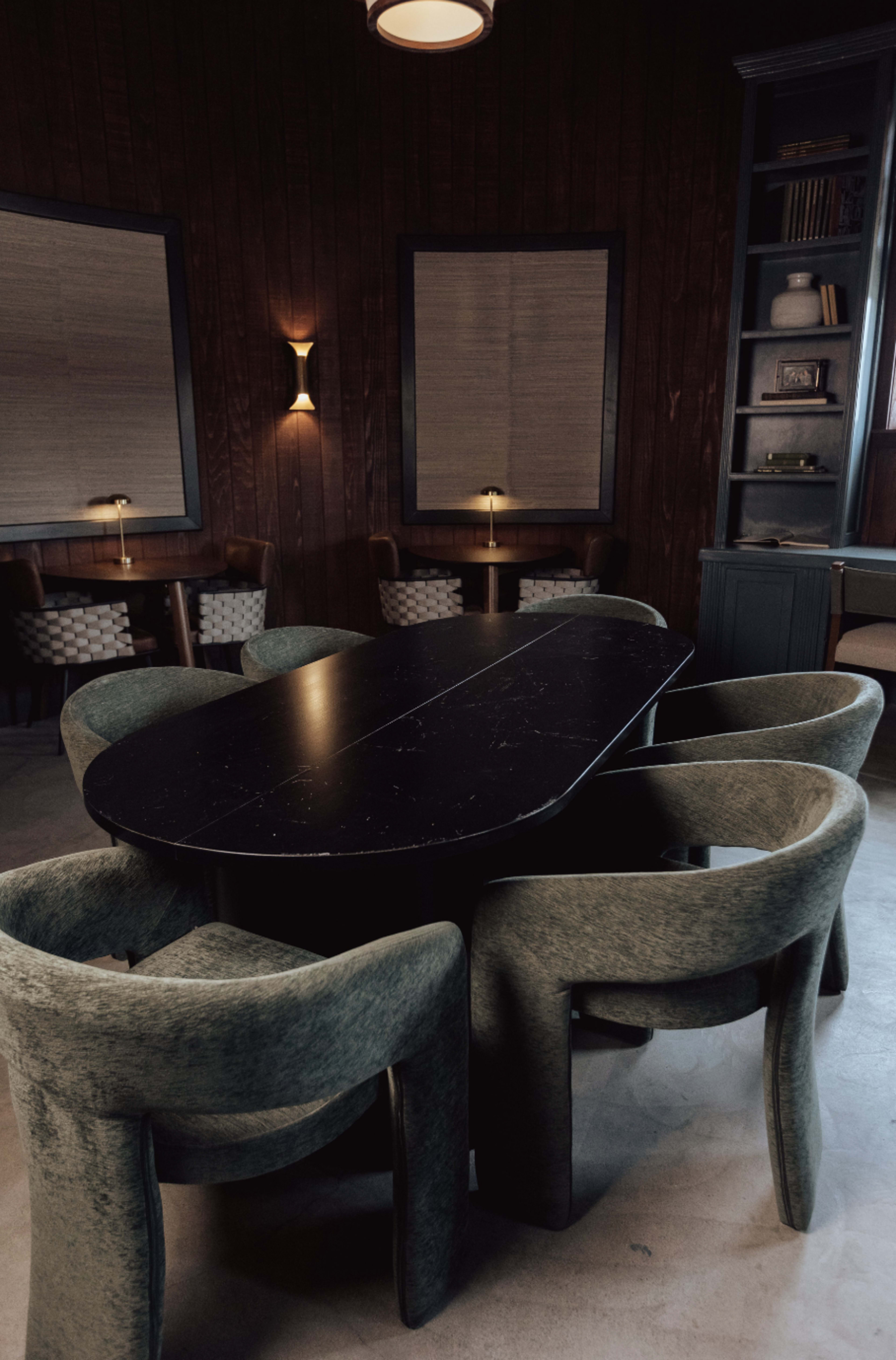 A large black oval table is surrounded by four green upholstered chairs in a dimly lit, wooden-paneled room.