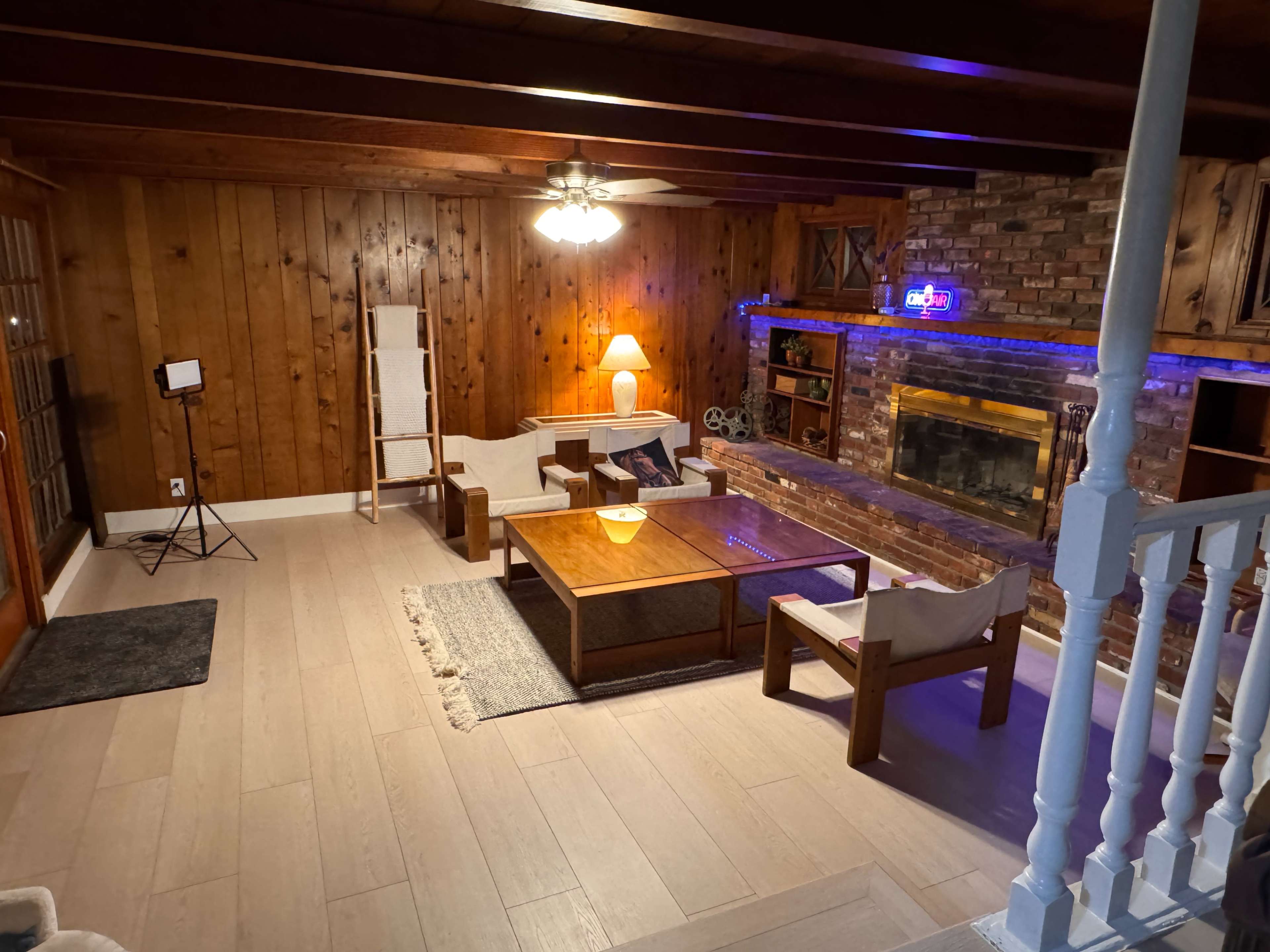 The image shows a well-furnished living room with wooden walls, a brick fireplace, a coffee table, and a warm lamp, creating a cozy atmosphere.