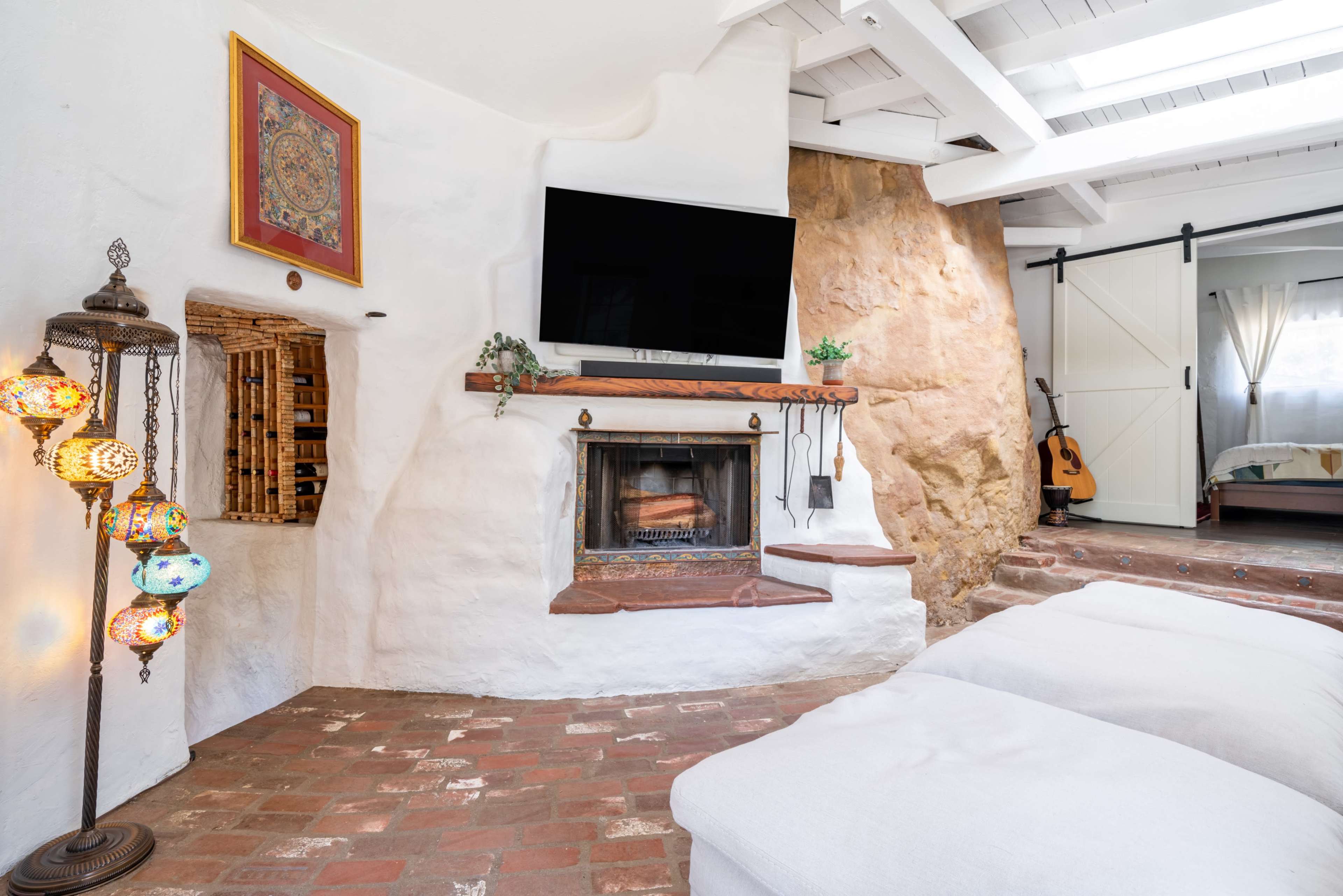 A cozy living room with a whitewashed wall featuring a framed artwork above a mantel, a modern flat-screen TV, a fireplace, decorative lanterns, and a guitar in the corner.