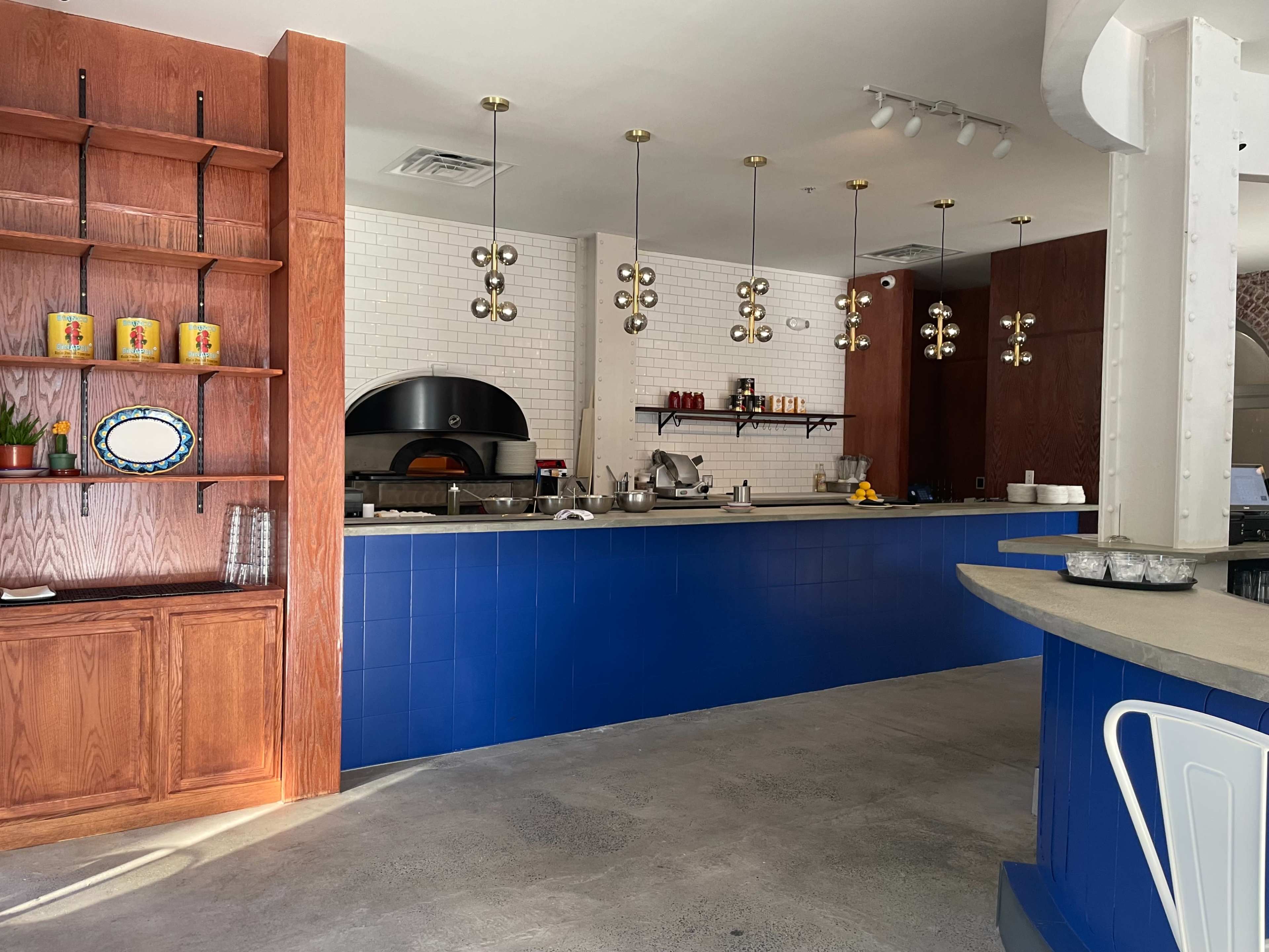 The image shows a modern restaurant interior with a blue tiled counter, wooden shelving, and decorative pendant lights above the bar area.