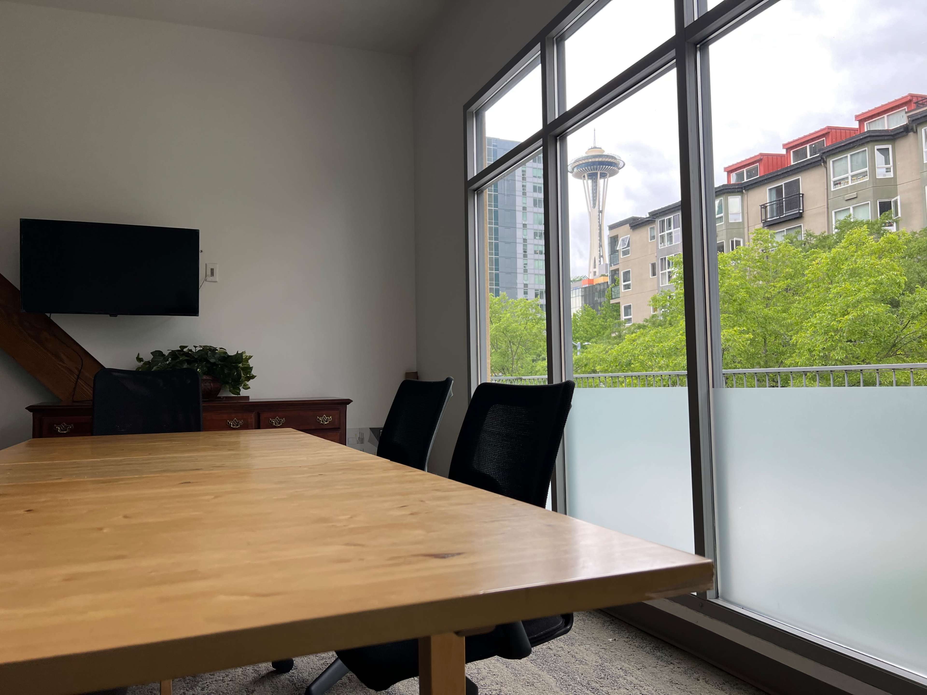 A wooden meeting table with black chairs is positioned in a conference room, overlooking green trees and the Seattle Space Needle through large windows.