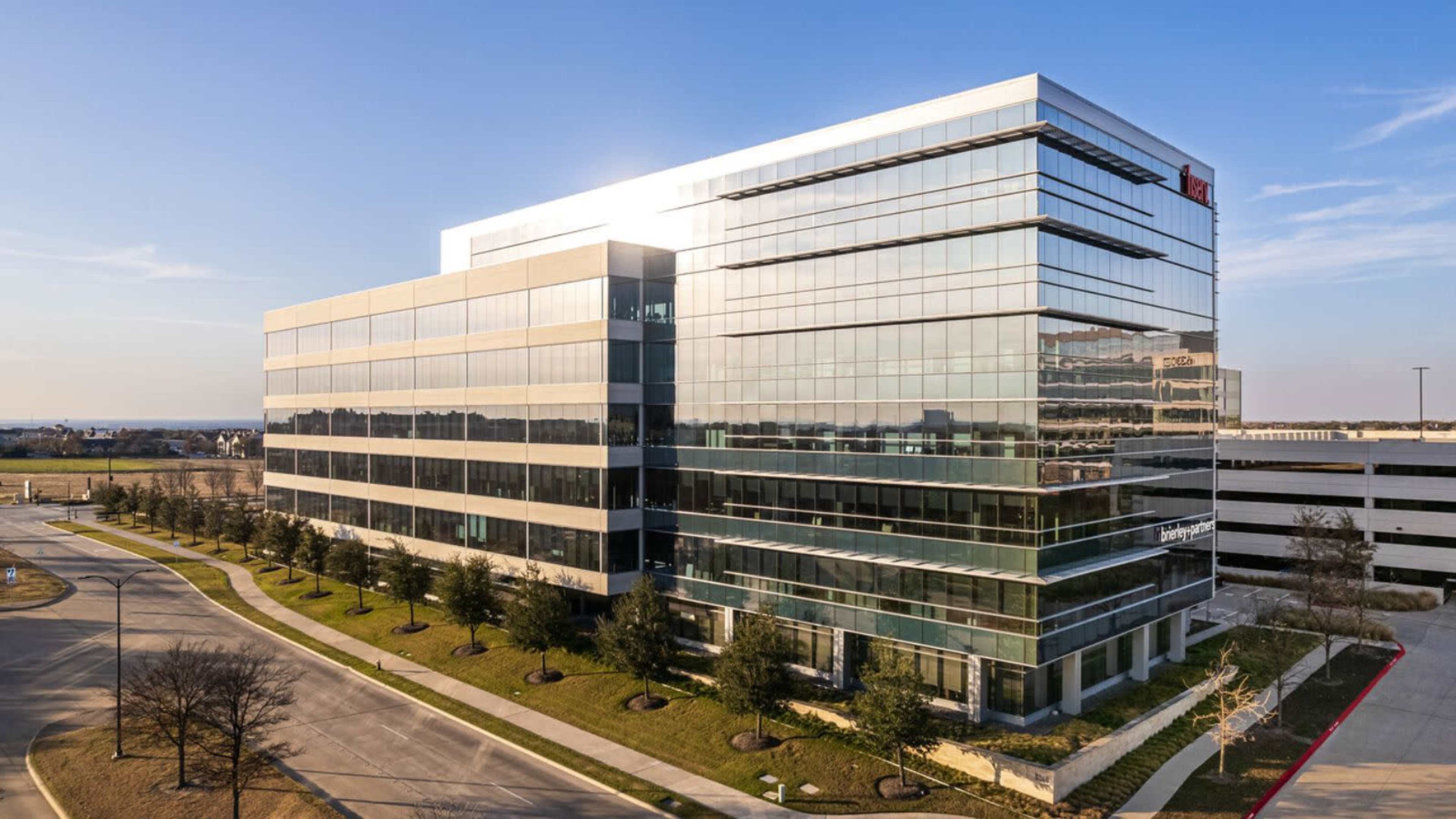 A modern glass office building with multiple stories and surrounding landscaped greenery is shown beside a parking structure.