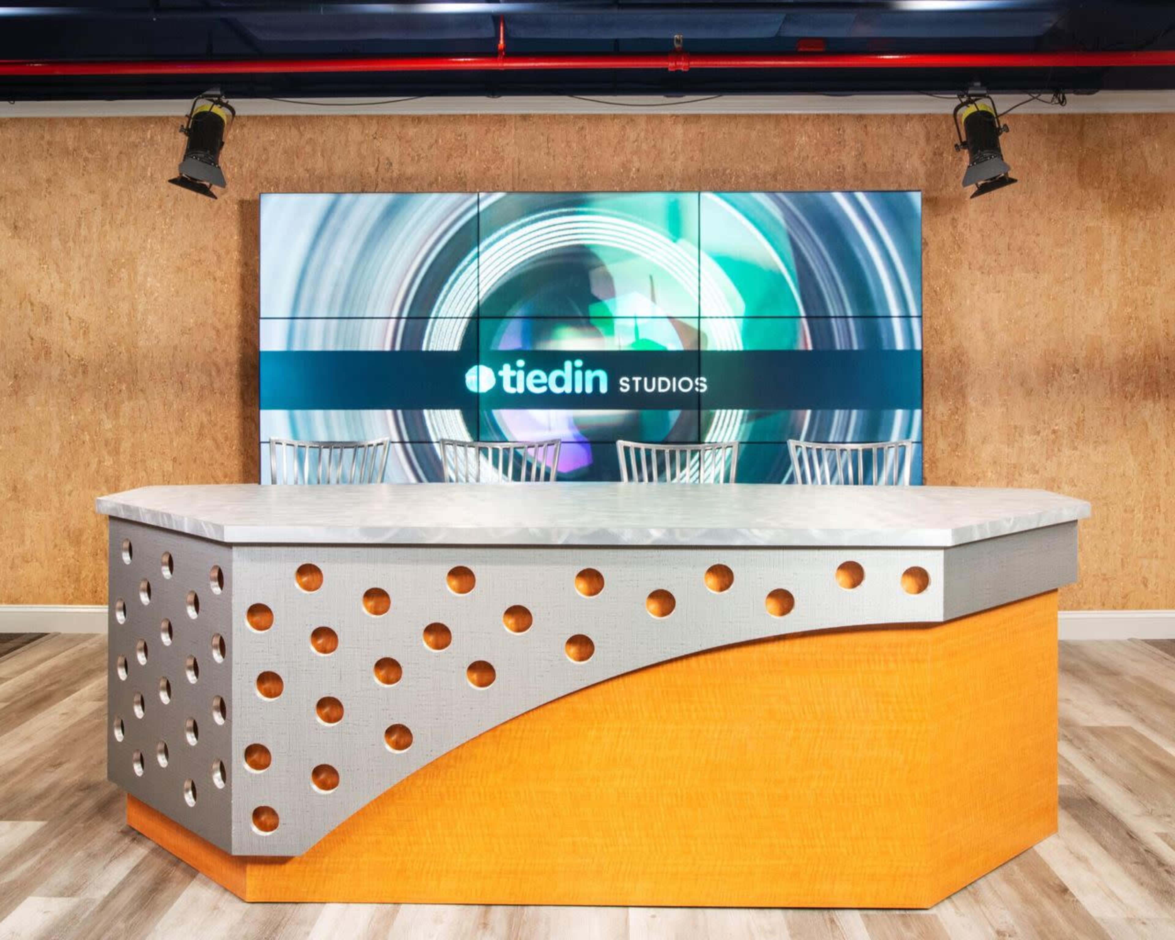 The image shows a modern studio setup with a curved gray and orange reception desk in front of a large digital display featuring the logo "tiedin STUDIOS."