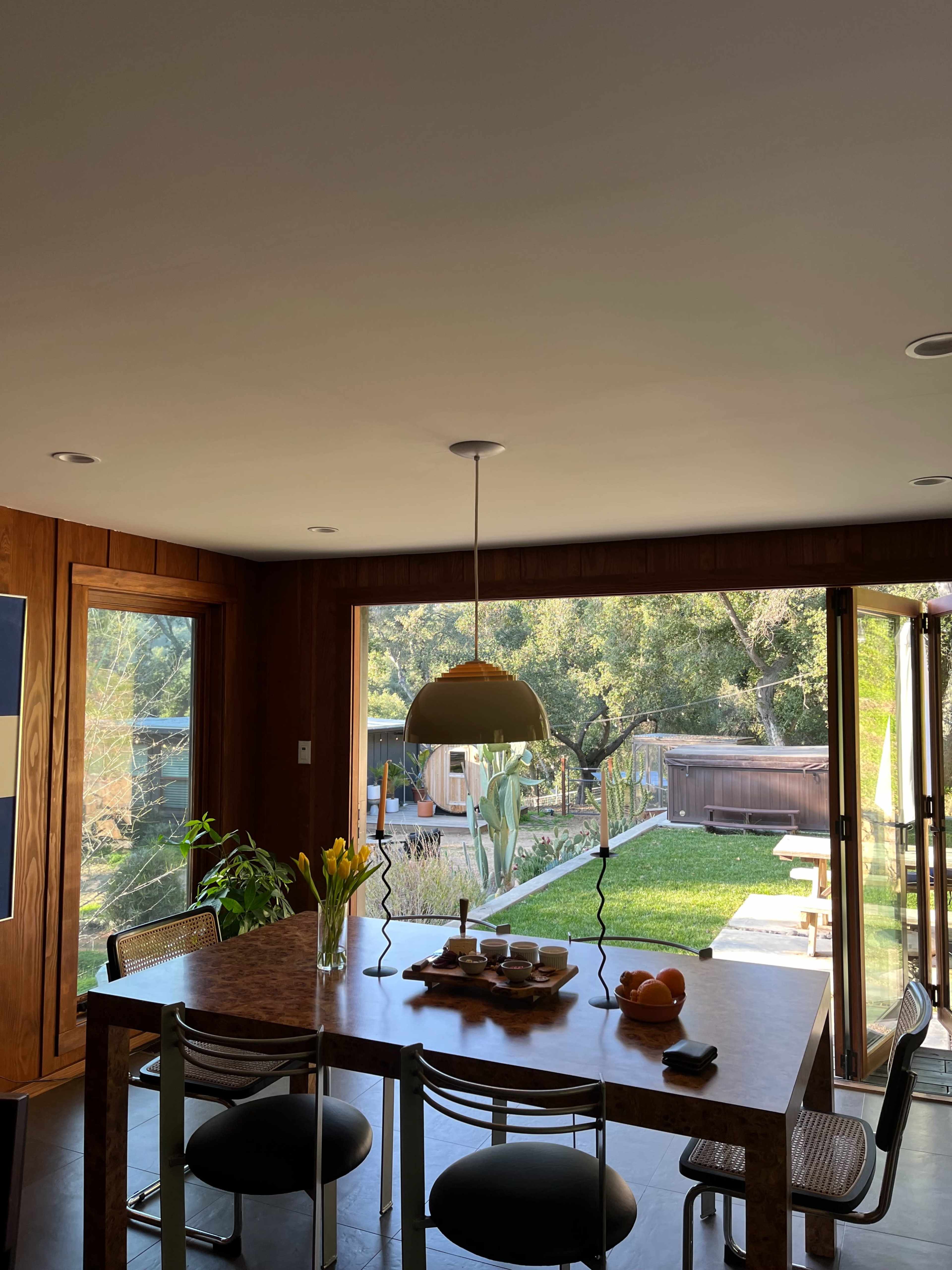 A dining area features a wooden table with a pendant light overhead, surrounded by chairs, and large sliding glass doors open to a green backyard.