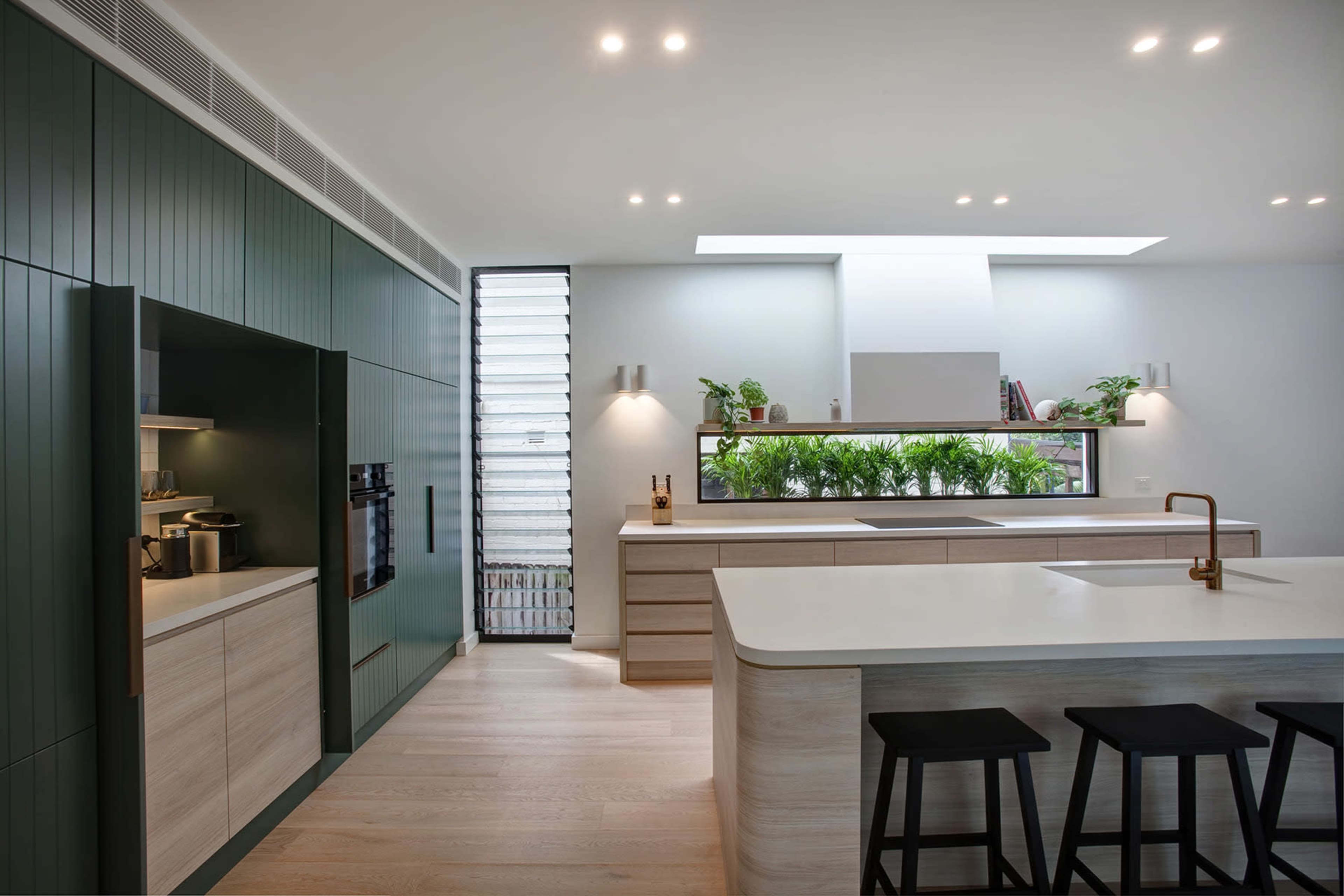 A modern kitchen featuring green cabinetry, a large island with a sink, and a window displaying indoor plants.