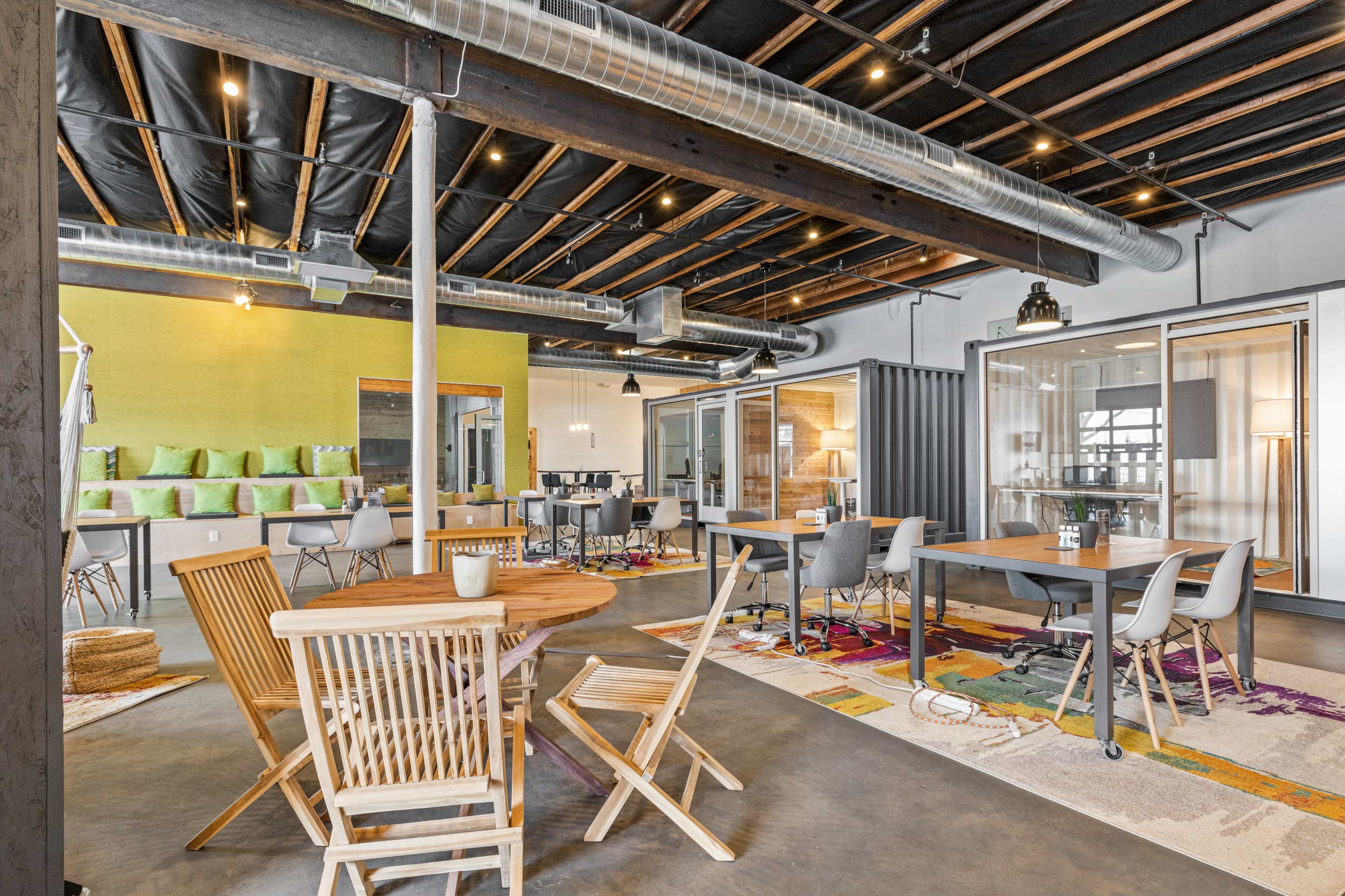 The image shows a modern workspace featuring various seating arrangements, wooden furniture, colorful rugs, and glass-walled conference rooms.