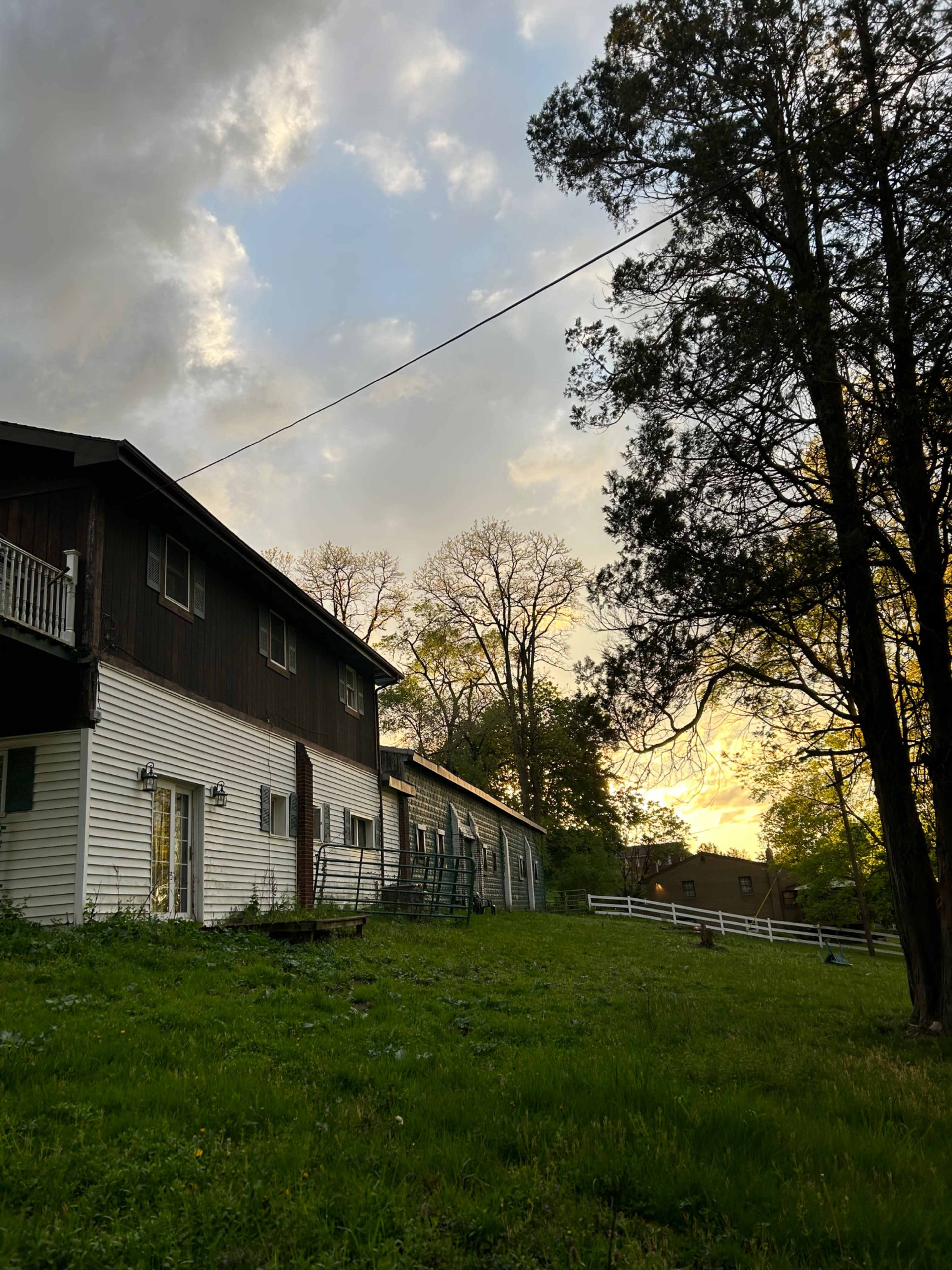 A large, two-story house with a dark wooden facade next to a grassy area and trees, under a cloudy sky during sunset.