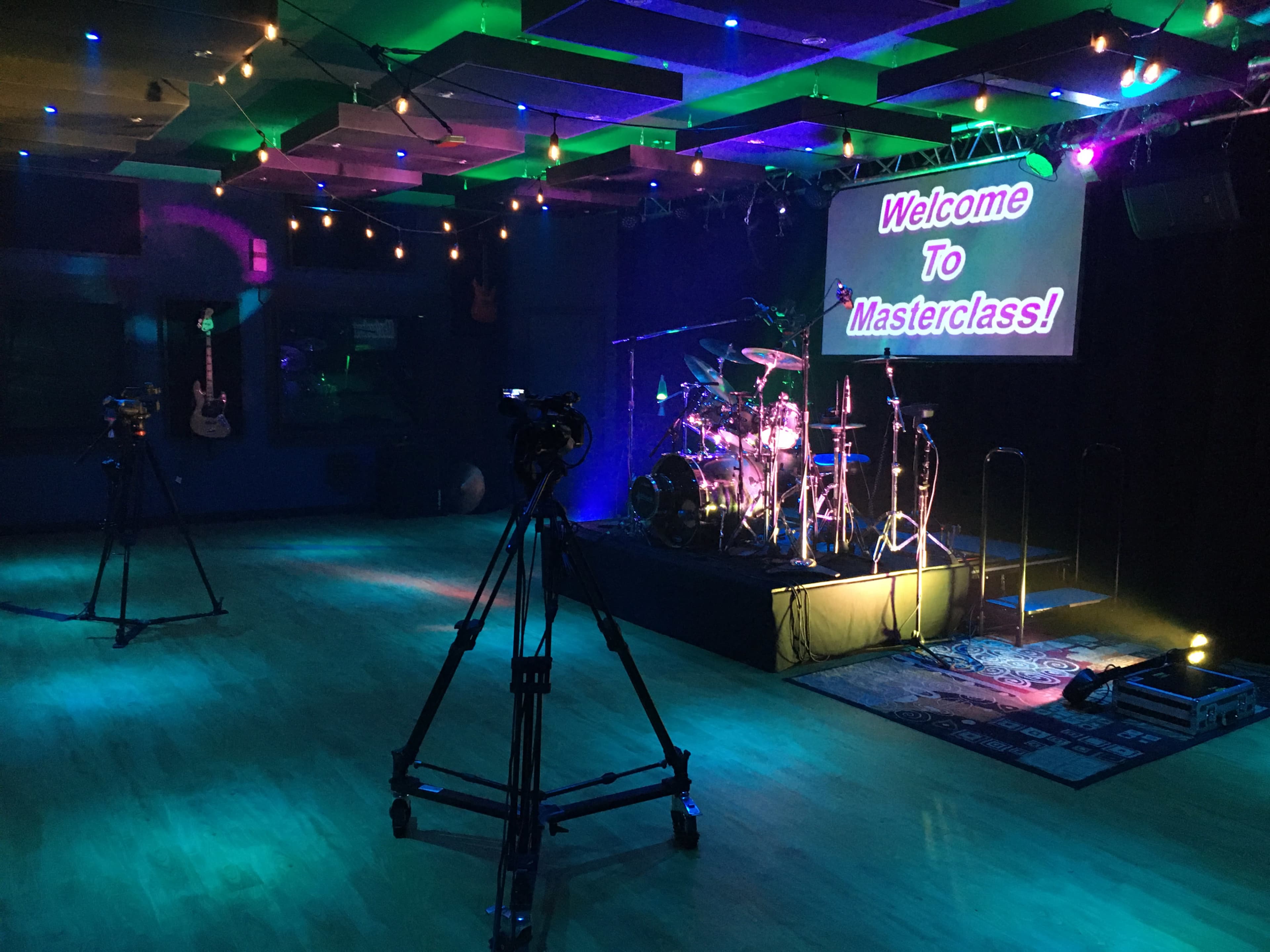 A stage set up for a masterclass, featuring a drum kit, cameras on stands, and a brightly lit sign that reads "Welcome To Masterclass."