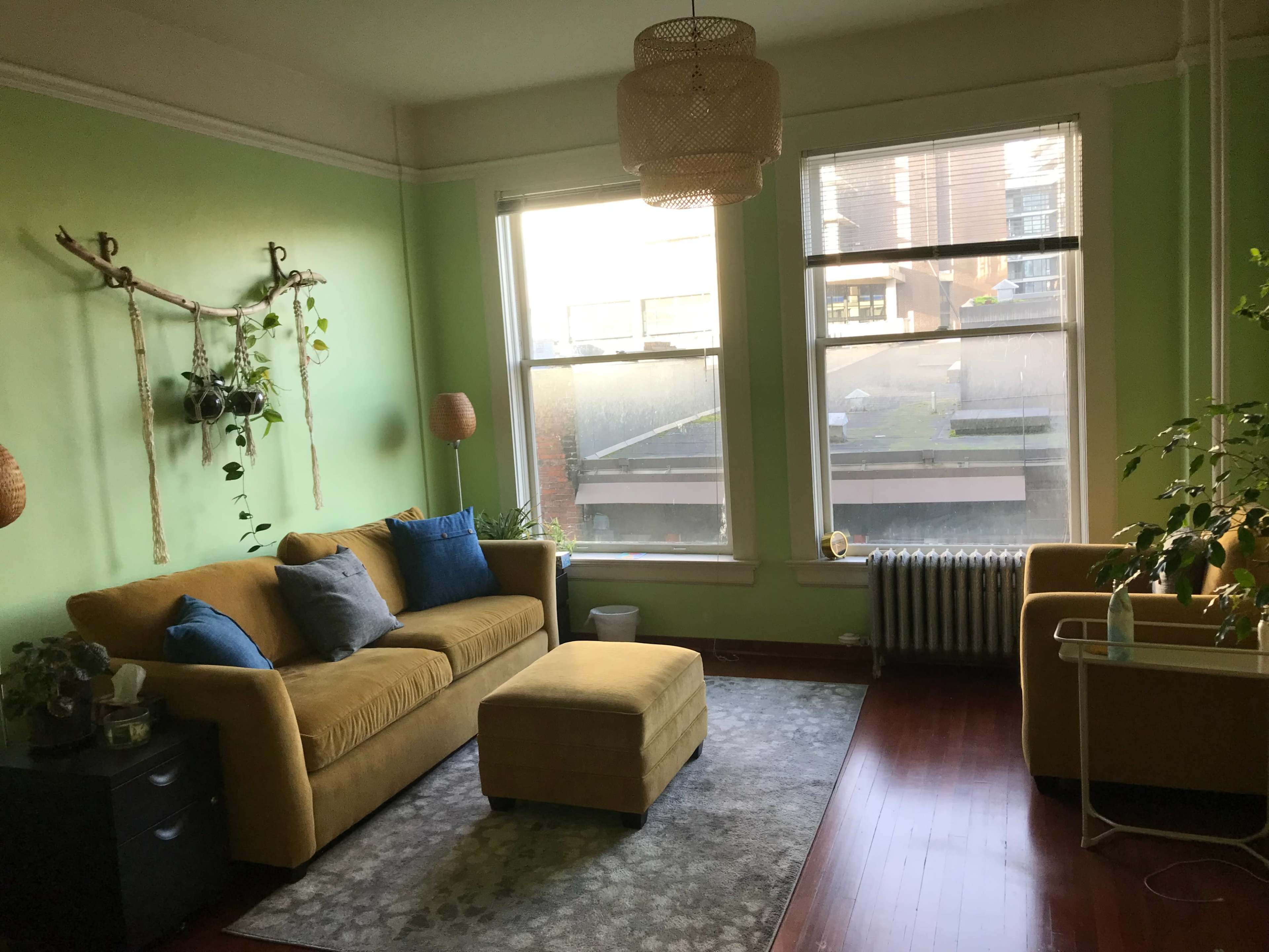 The image shows a living room with green walls, a mustard yellow sofa, an ottoman, and large windows allowing natural light to fill the space.