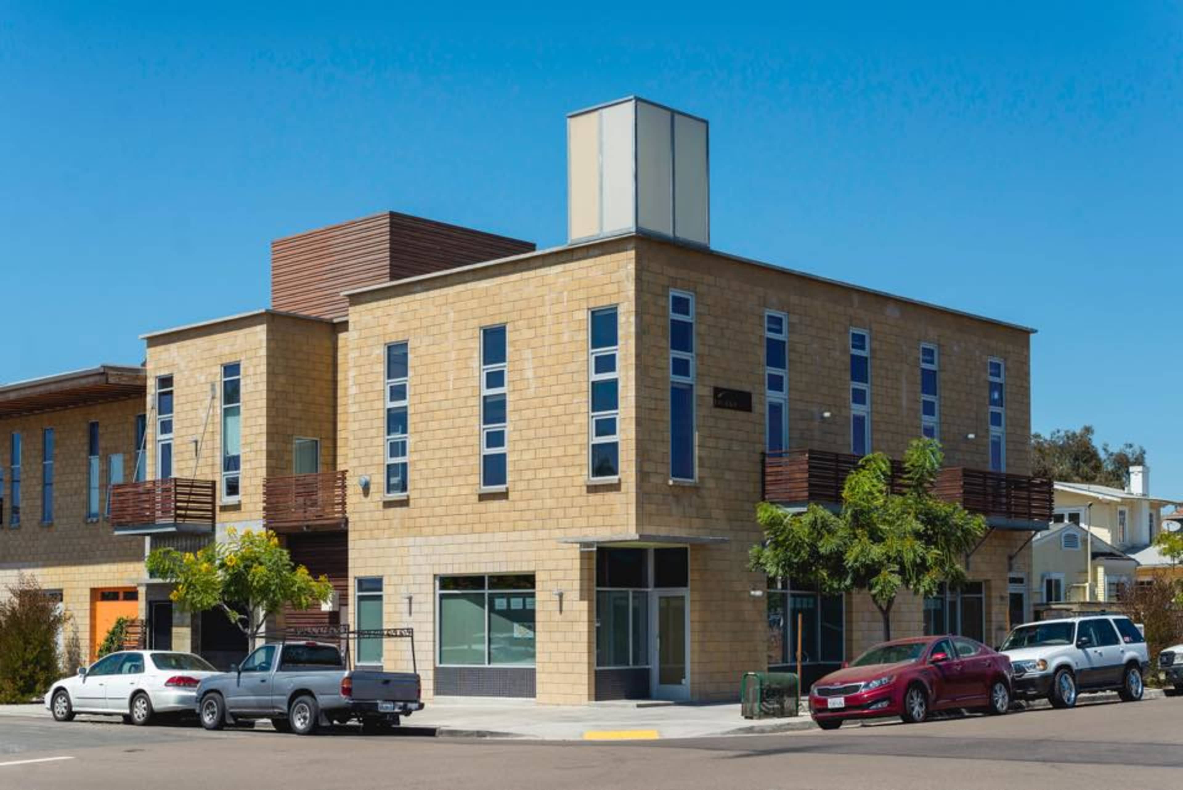 A modern two-story building with a stone facade and large windows, situated at a street corner with parked vehicles in front.