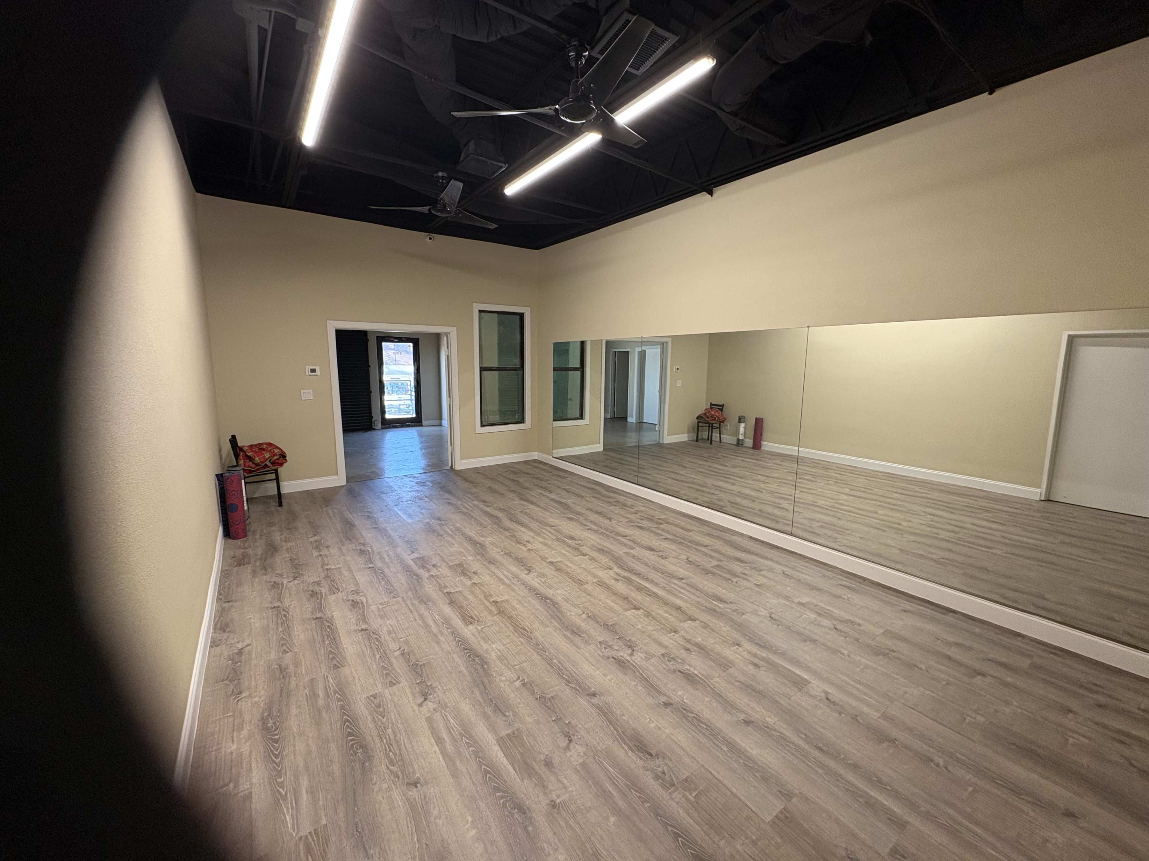 The image shows a spacious, empty room with wooden flooring, a large mirror on one wall, and a ceiling fan.