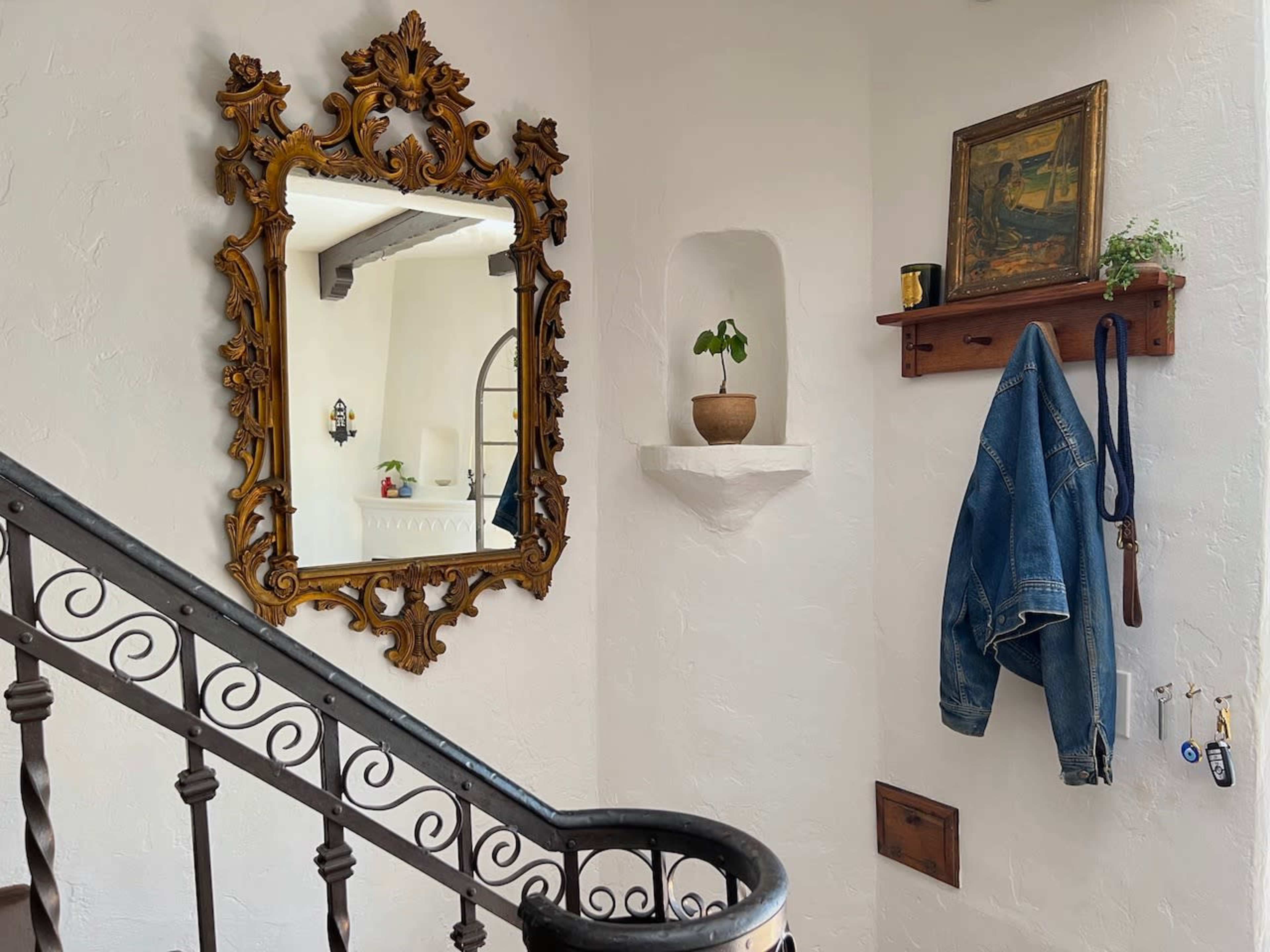 The image shows a staircase with ornate railing, a large decorative mirror on the wall, a small plant on a shelf, and a blue denim jacket hanging beside a wooden shelf containing a painting.