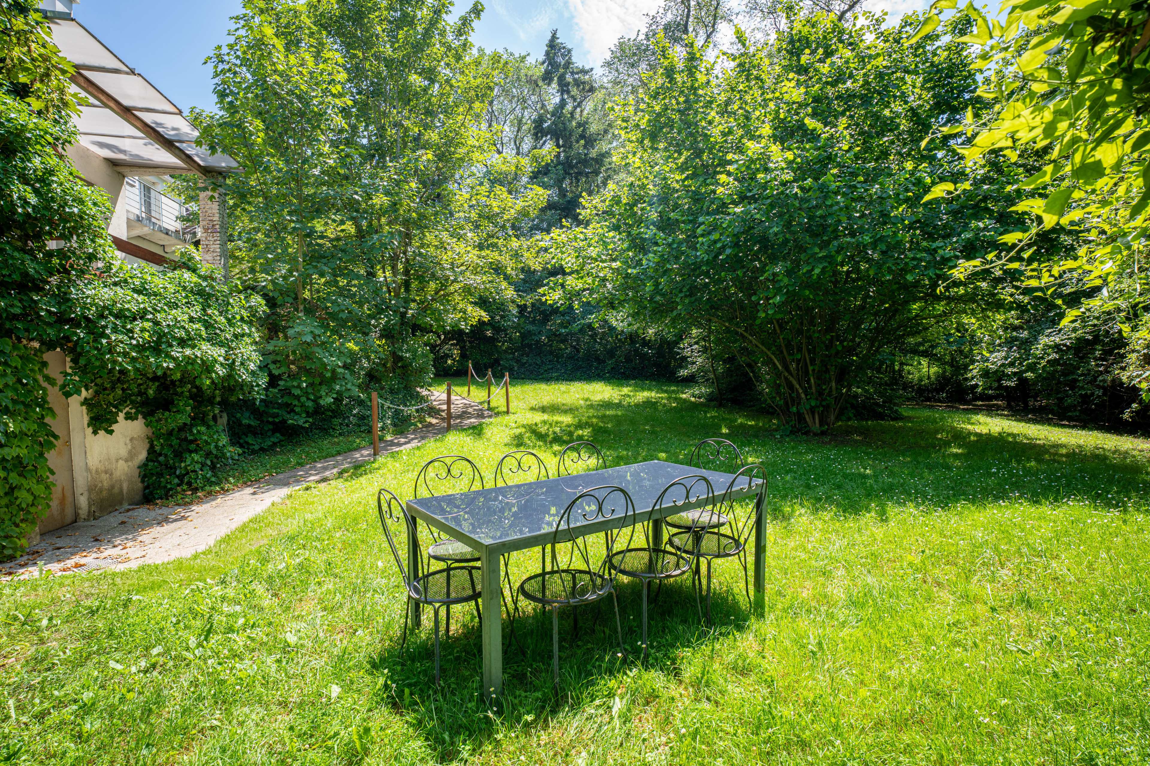 A rectangular table with six chairs is positioned on a grassy area surrounded by trees and shrubs.