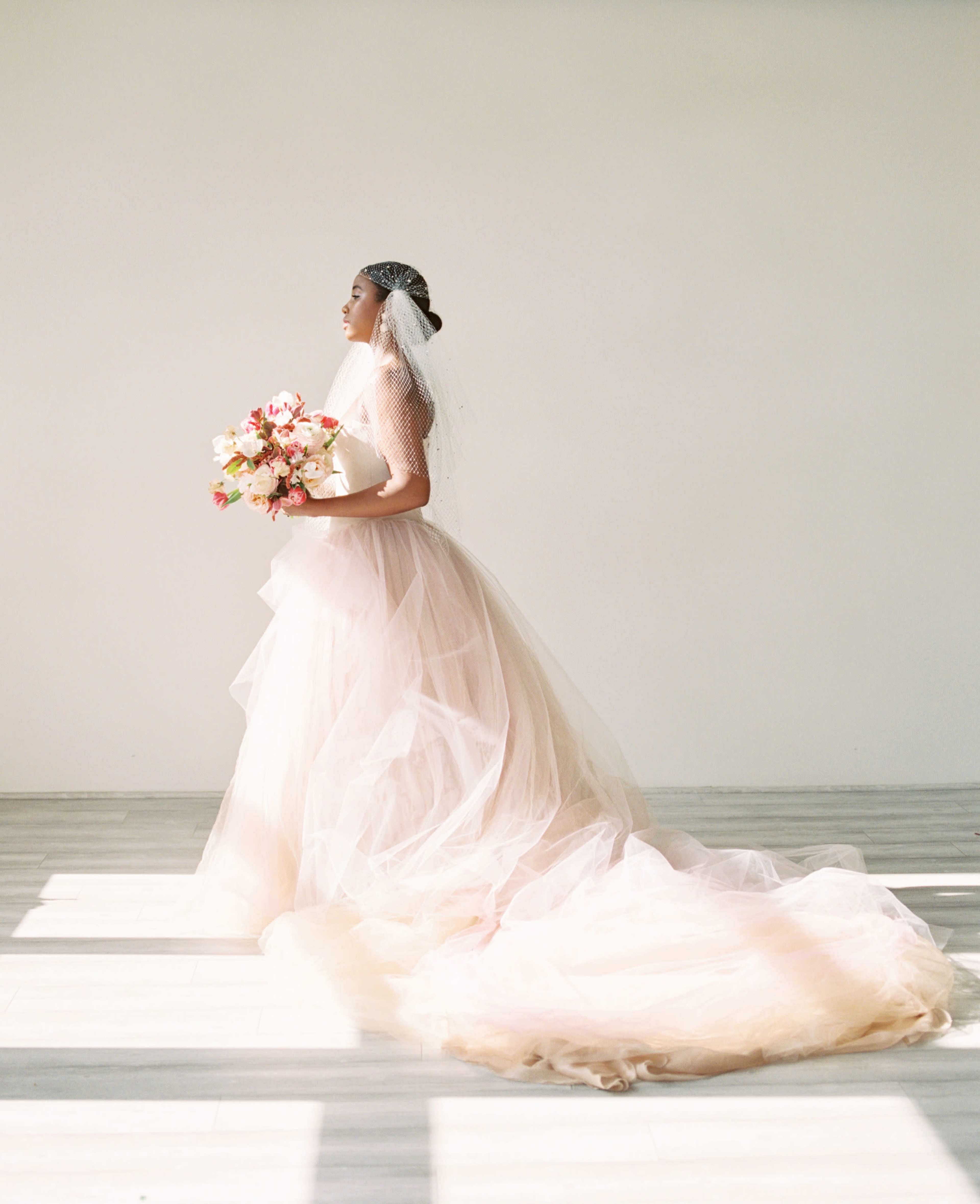 A bride in a flowing, tulle wedding gown holds a bouquet while standing against a plain wall.