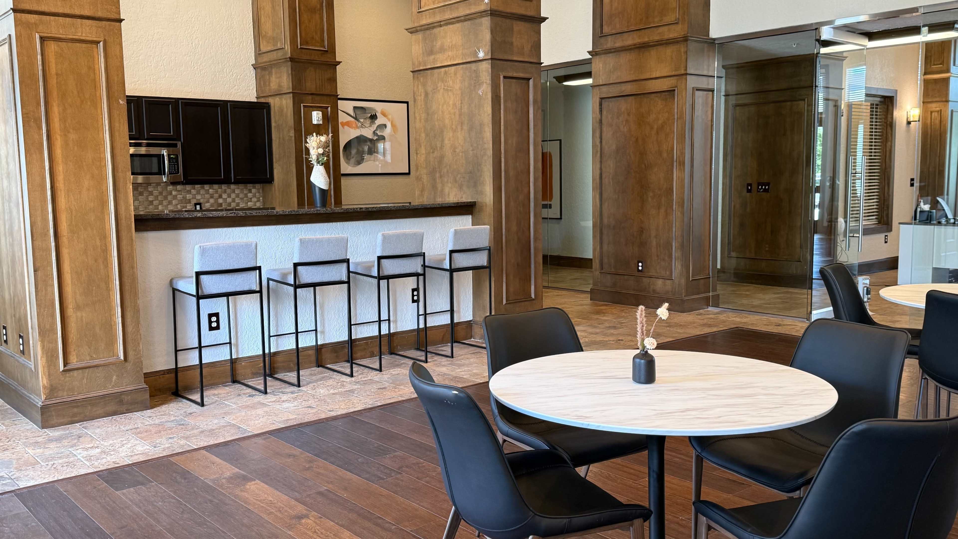 The image shows a modern communal space with a kitchenette featuring bar stools, a round dining table, and wooden pillars.