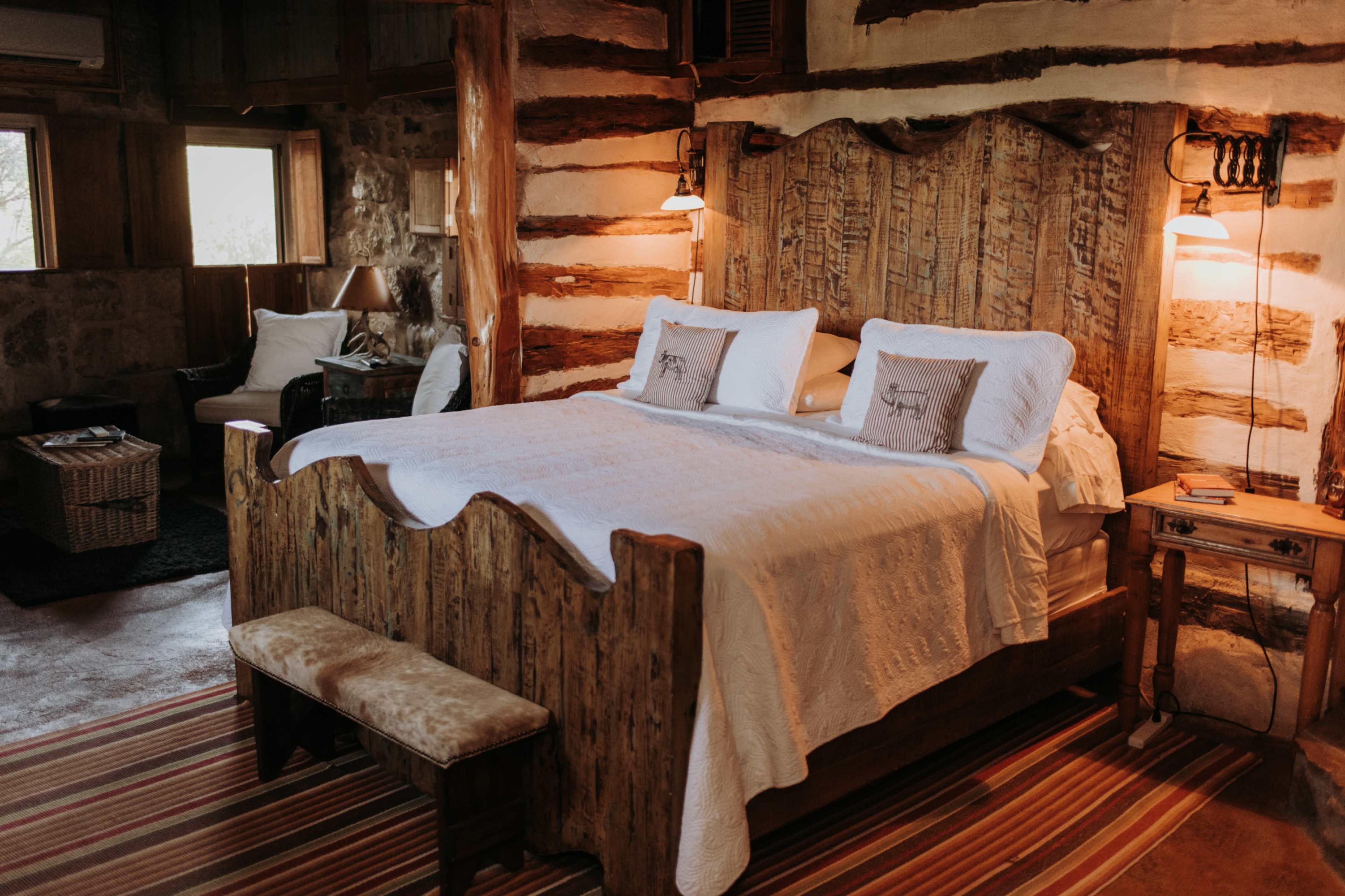 The image depicts a rustic bedroom featuring a wooden bed with a headboard, white bedding, and lamps on either side, set in a room with exposed log walls and a striped rug.